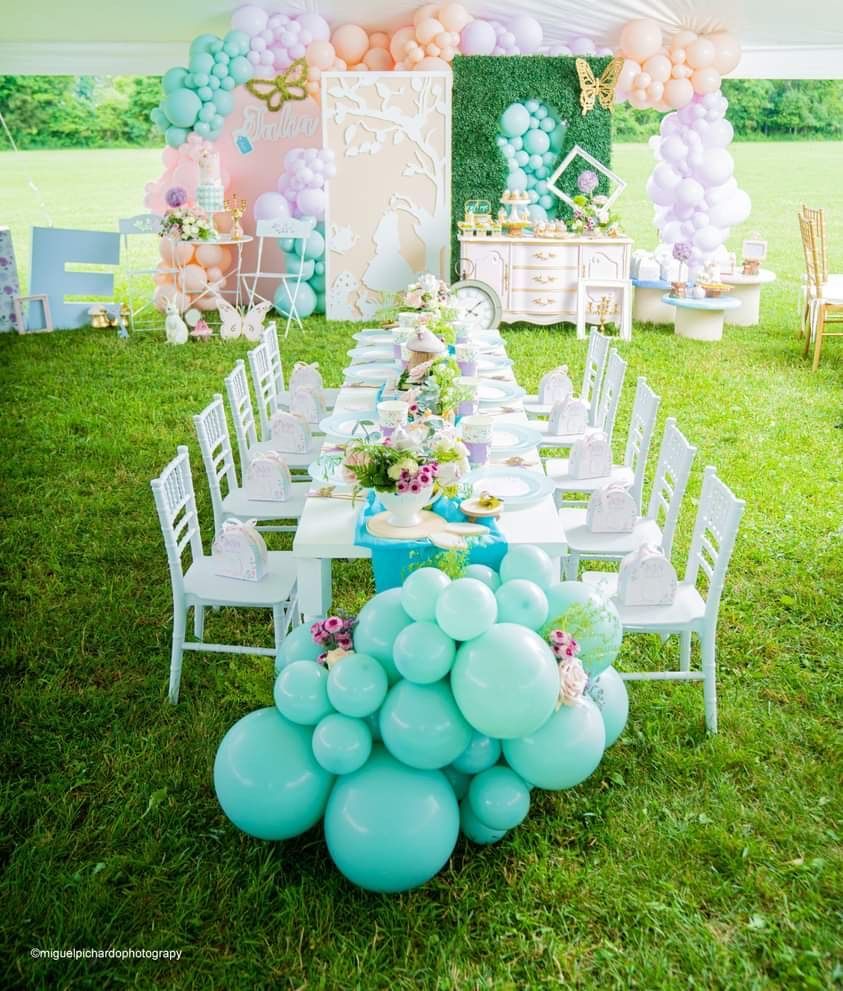 Pastel balloon-decorated outdoor party table on a grassy lawn with white chairs and floral decor