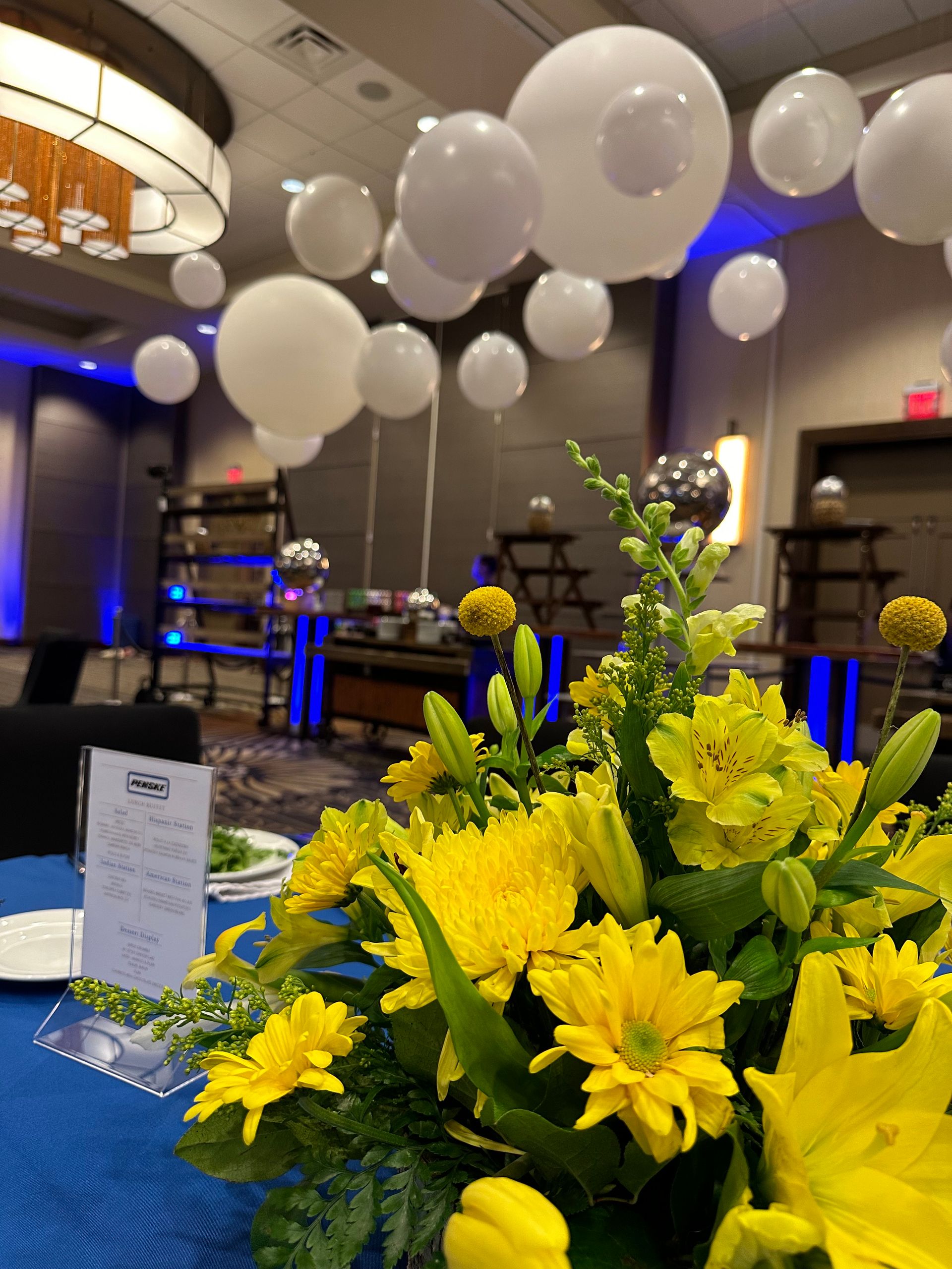 Yellow flowers on a blue table at a banquet hall with white balloons and blue-lit tables in the background