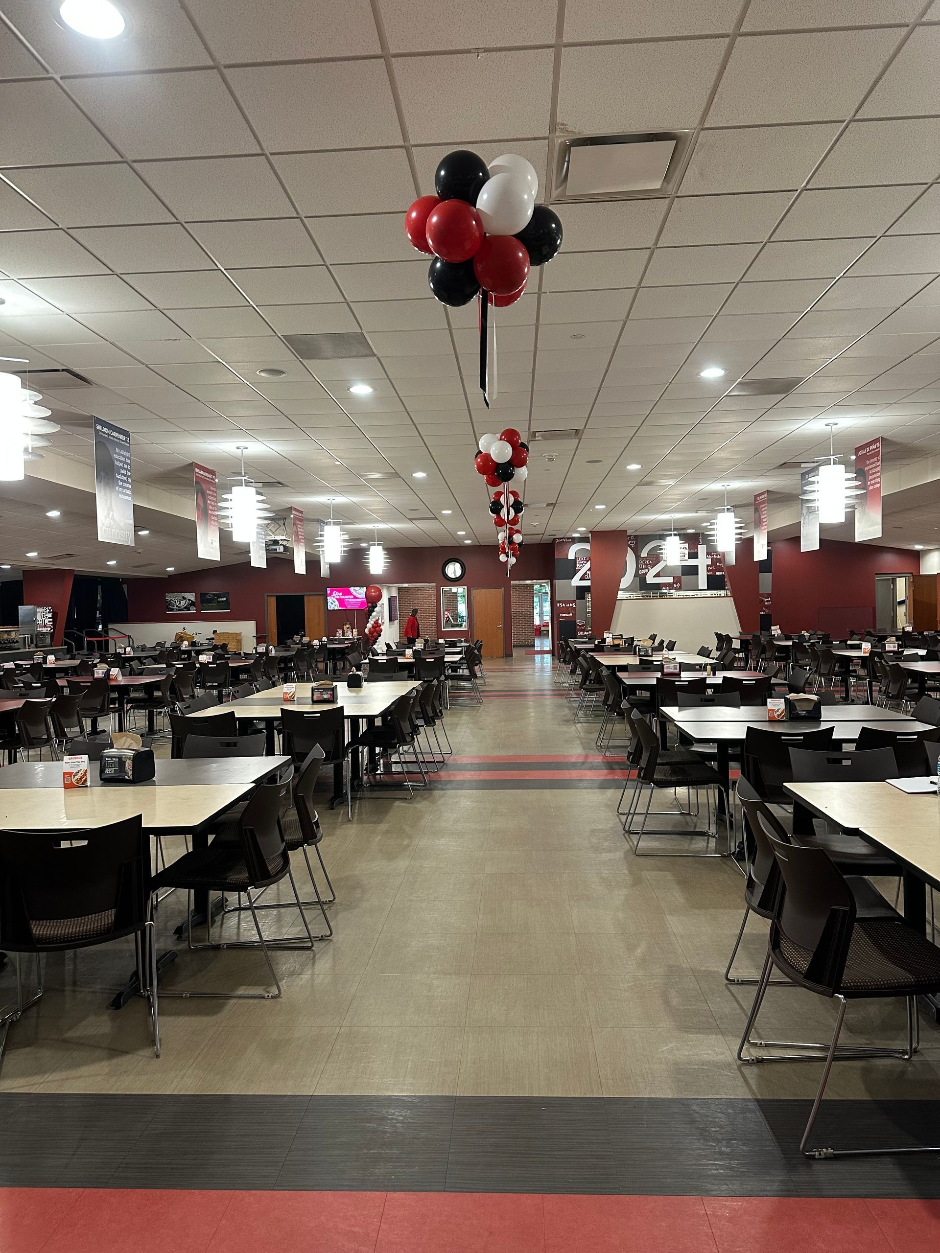 Decorated banquet hall with rows of tables and chairs, black-white-red balloons hanging from the ceiling.