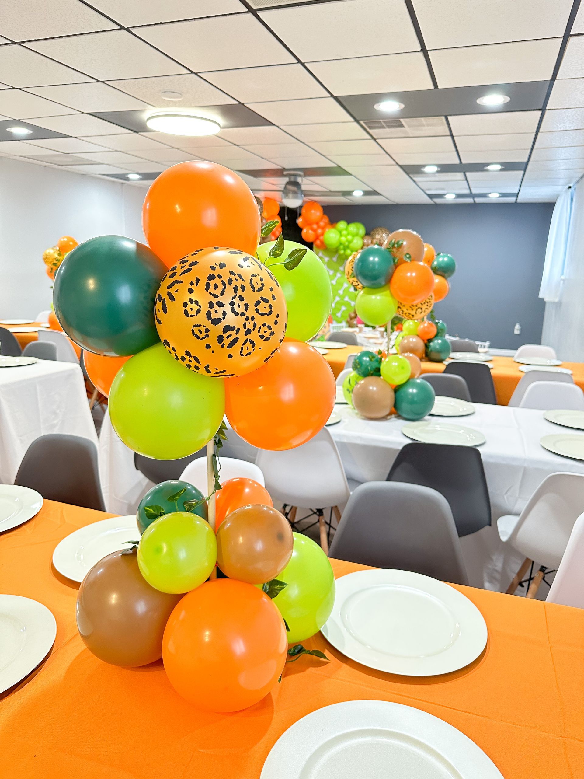 Orange and green balloon centerpieces on tables in a decorated banquet room with white plates and chairs