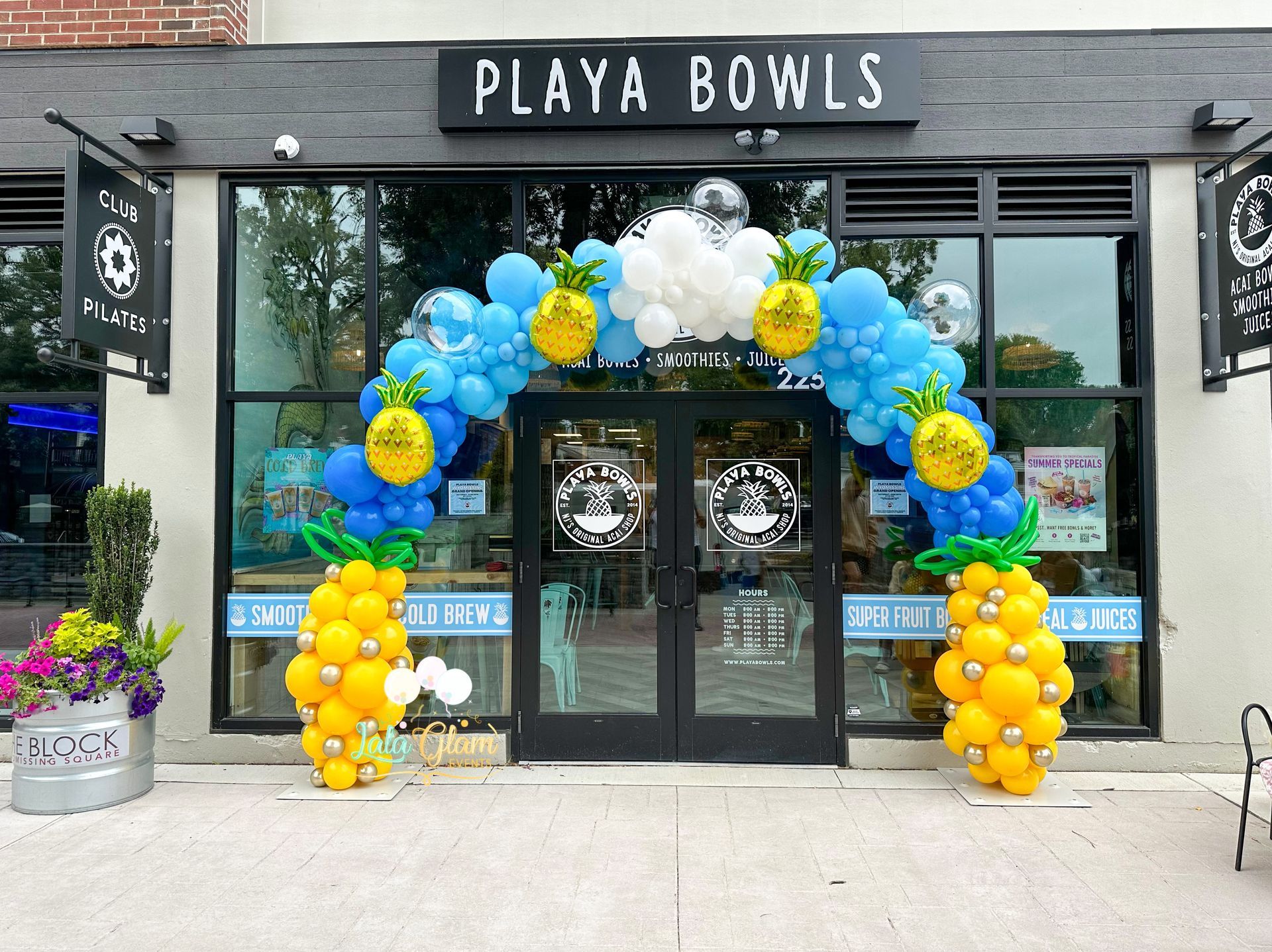 Exterior of a Playa Bowls storefront with blue and yellow balloon arch and pineapple decorations.