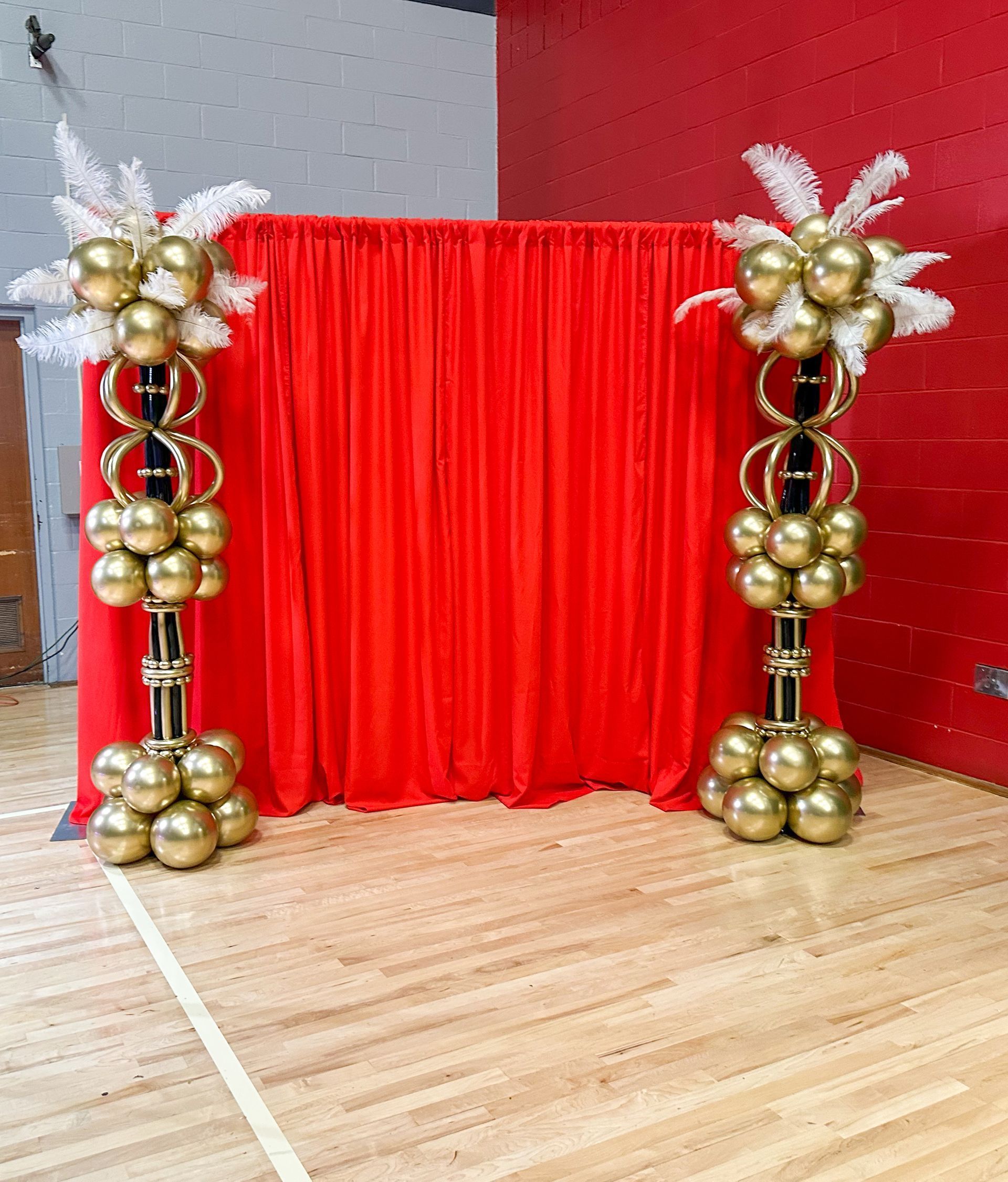 Two gold balloon columns flank a red curtain backdrop on a wooden floor. Each column has white feather accents.