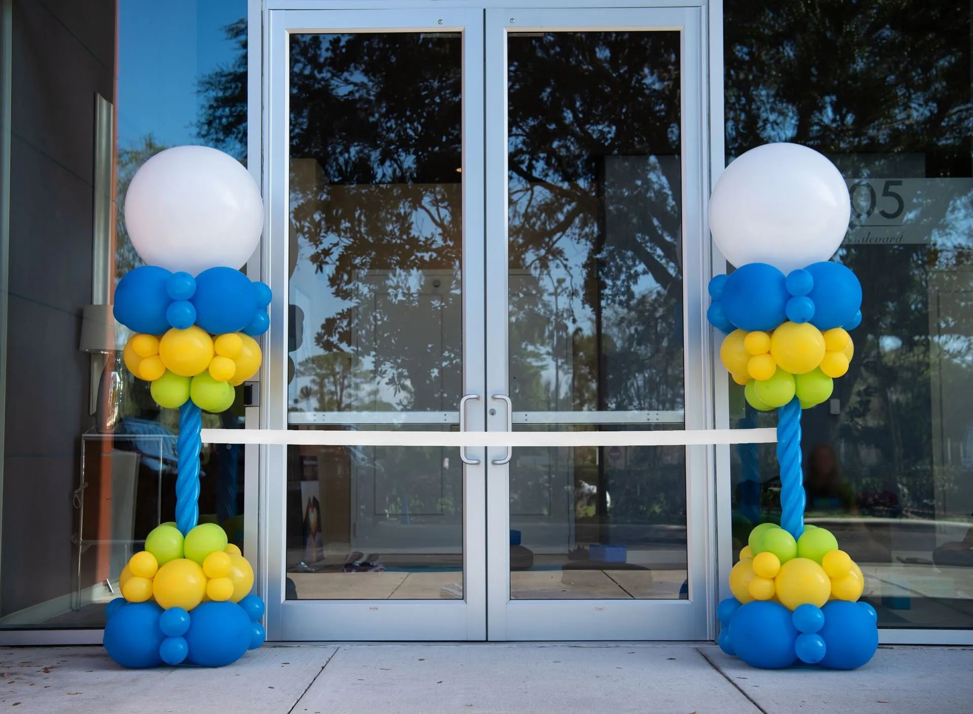 Two balloon columns flank double glass doors, a ribbon stretched between. Columns: blue, yellow, and white balloons.