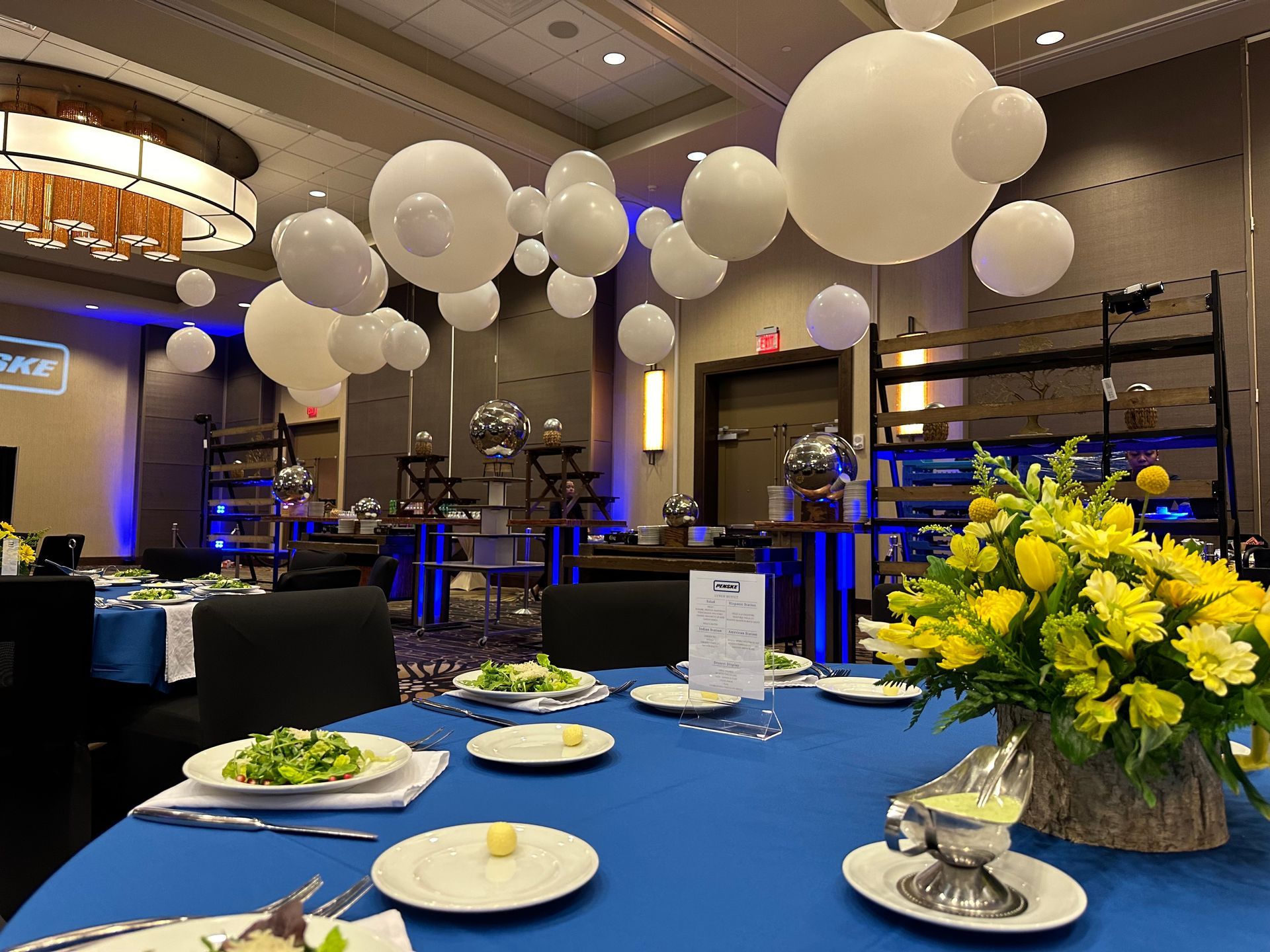 Banquet hall table with blue cloth, plates, and yellow floral centerpieces under white balloons