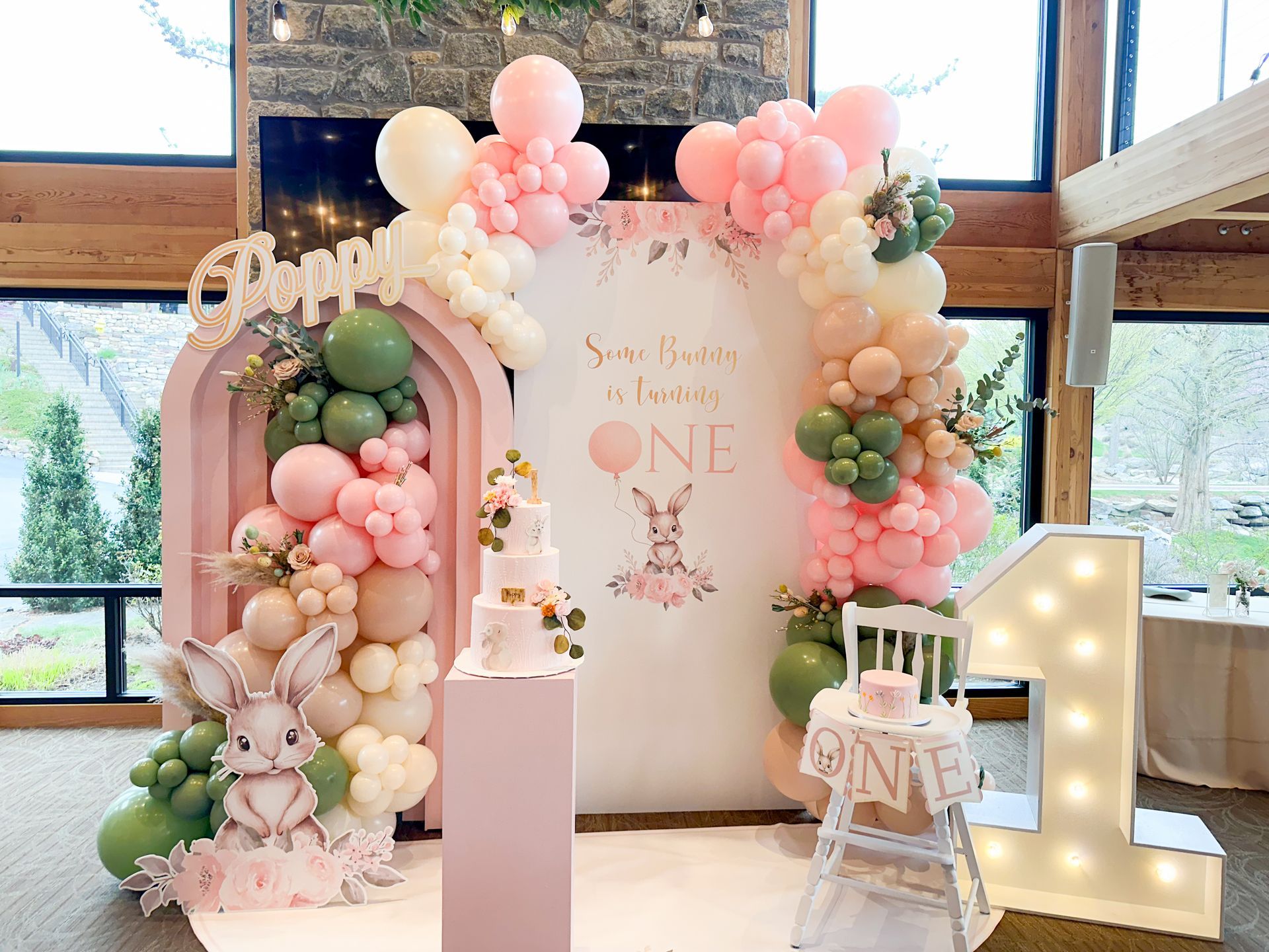 First birthday party with pink and green balloon arch, bunny decor, and cake.