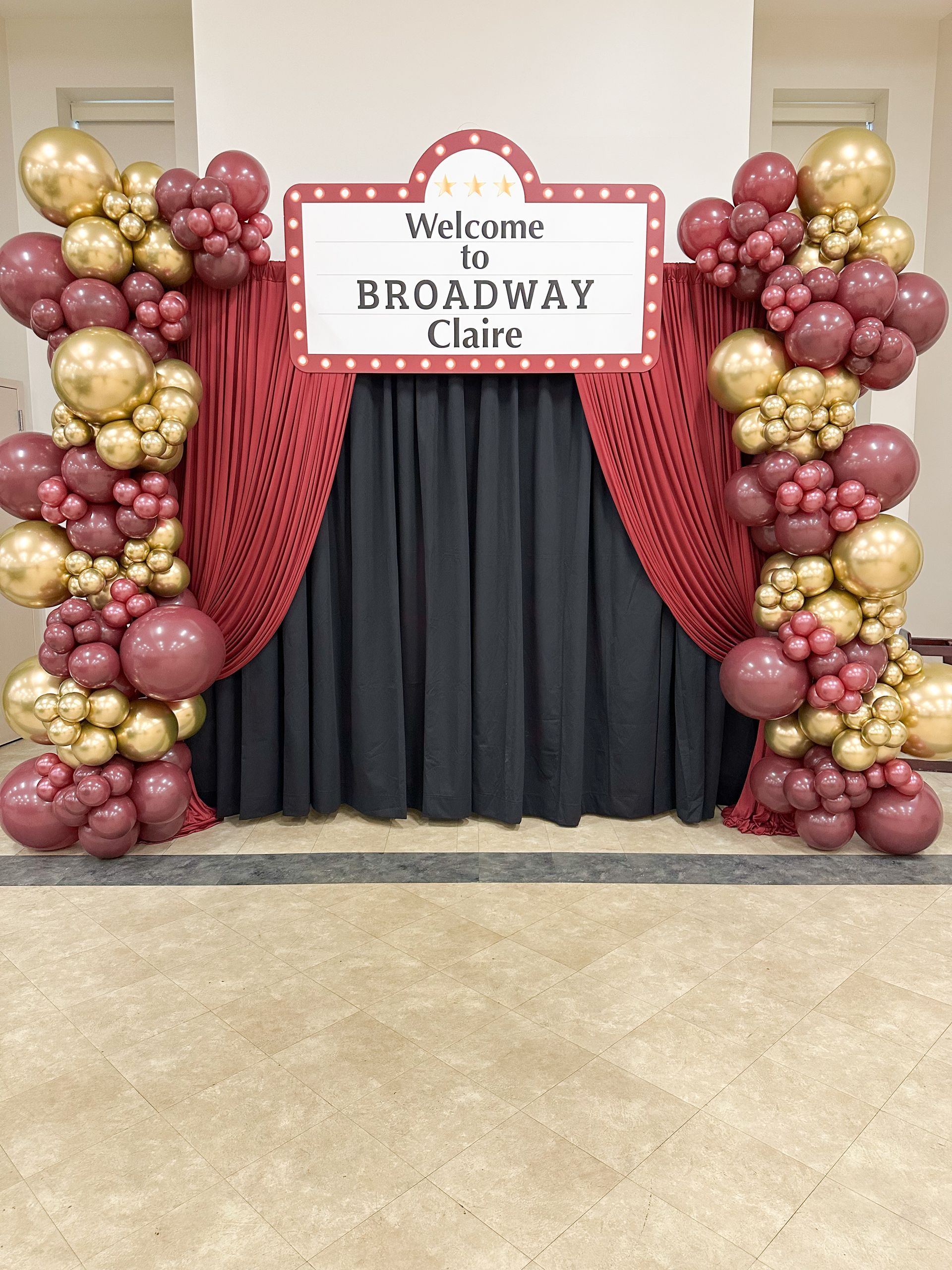 Broadway-themed party backdrop with maroon, gold balloons, a red curtain, and a sign that says,