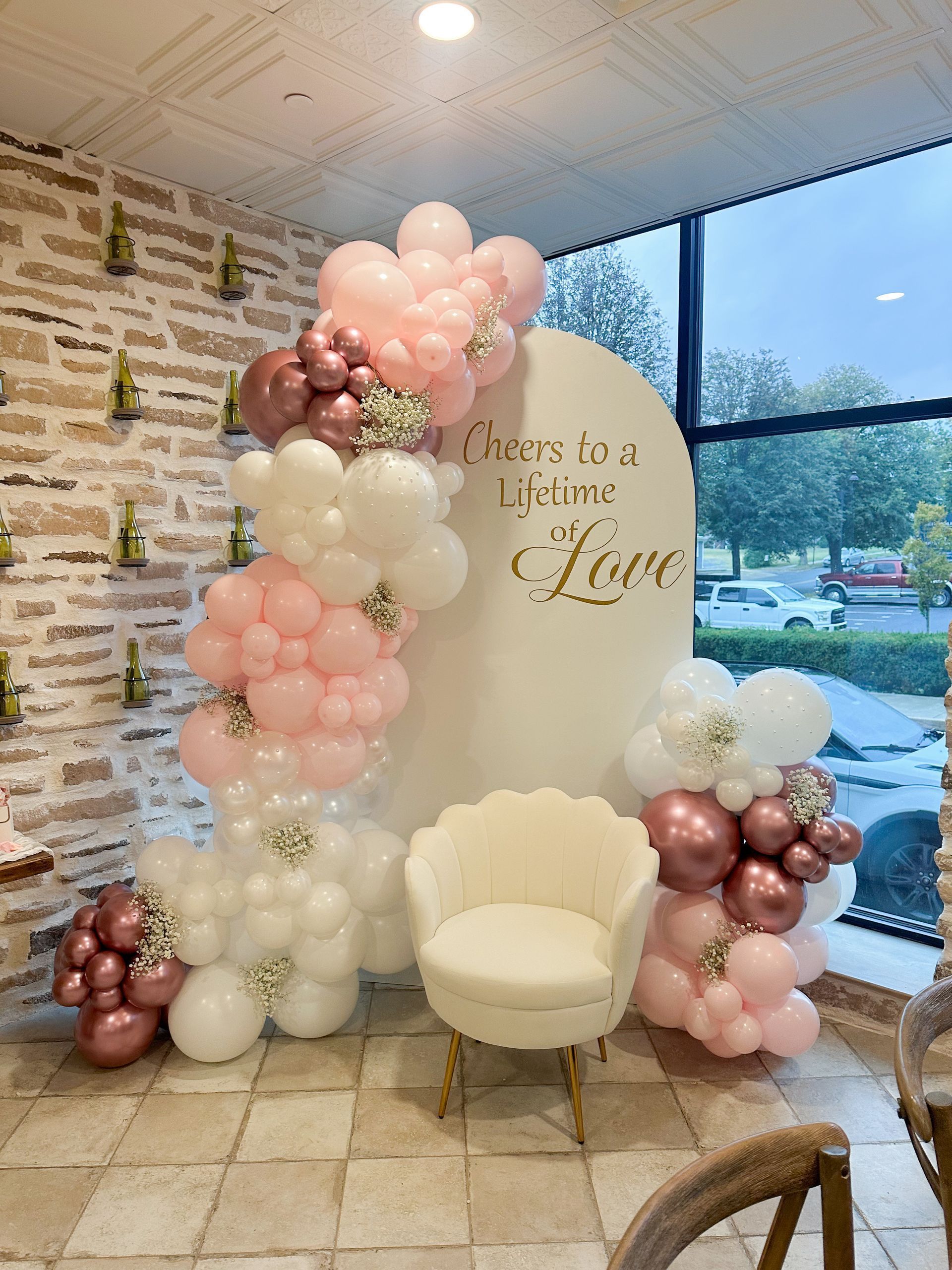 Balloon arch with a sign, chair, and large window. Pink, white, and bronze balloons. Gold text on the sign.