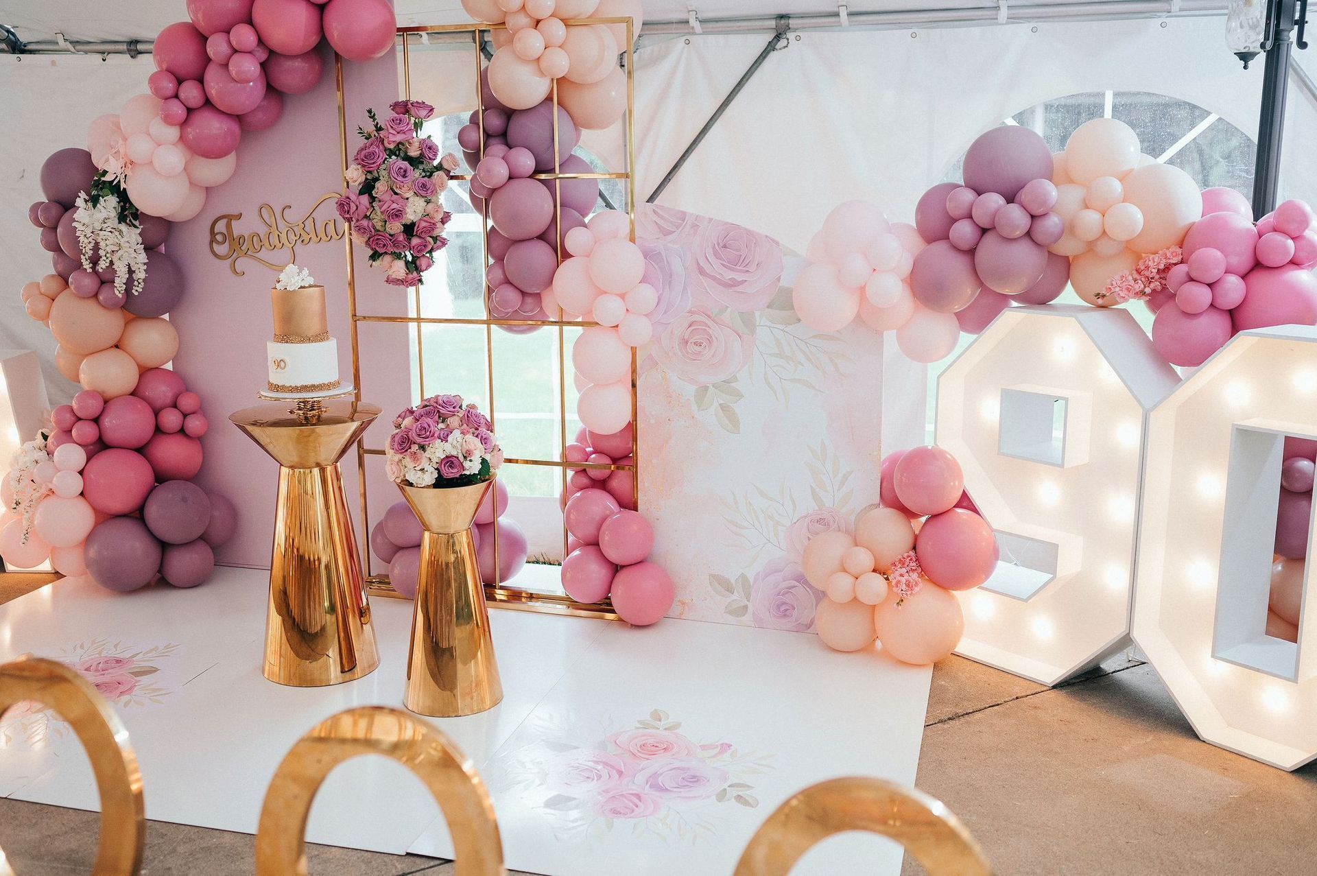 Pink and gold balloon-decorated party backdrop with illuminated letters and cake stands