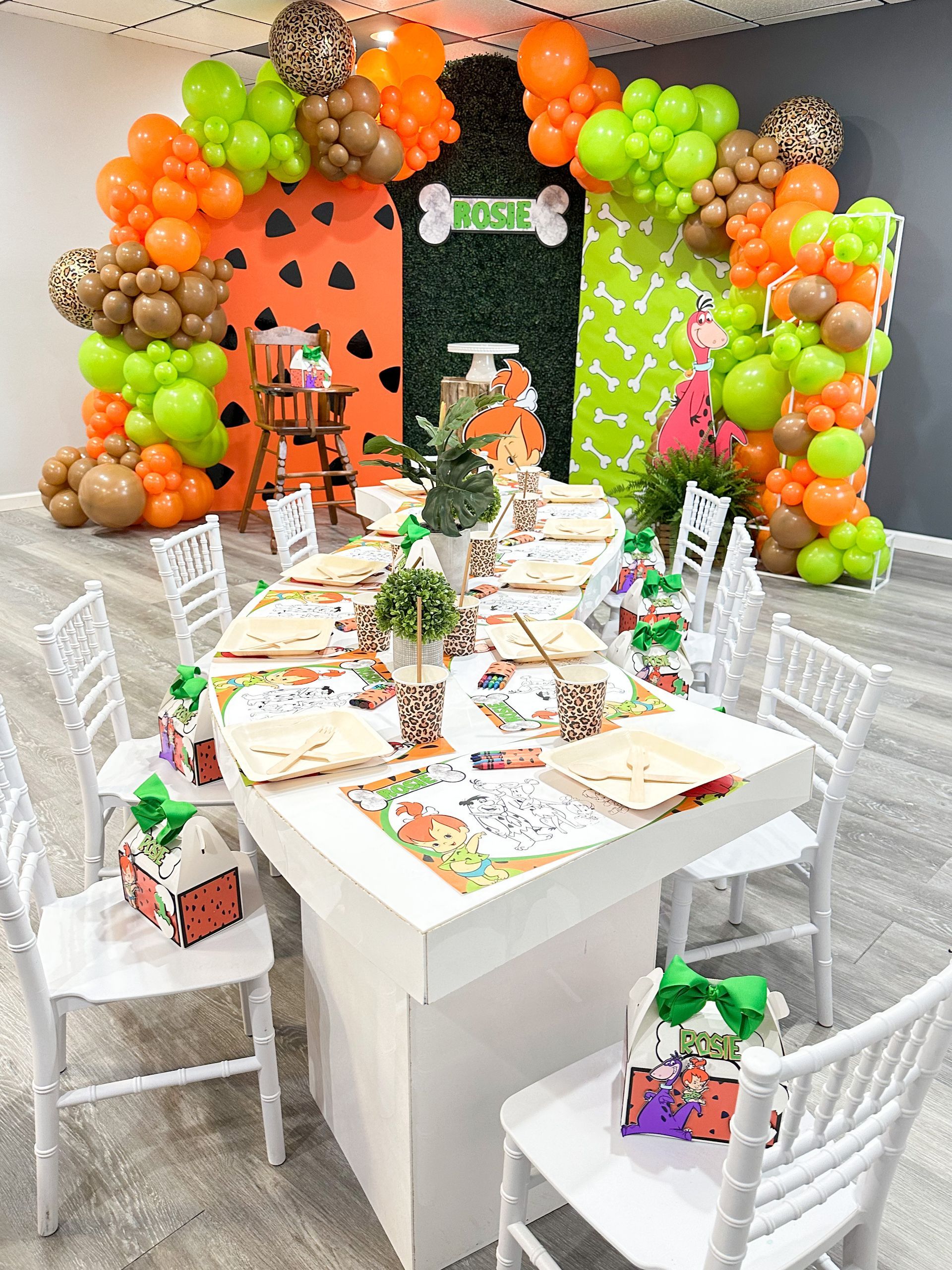Party table set with decorations; green, orange, brown balloons arch. White chairs, table with coloring pages, gifts.