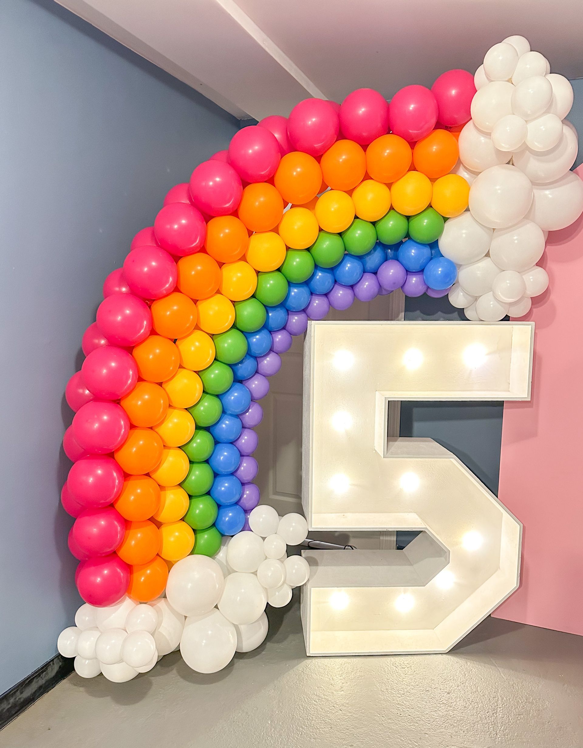 Rainbow-colored balloon arch with white cloud accents and a lighted number 