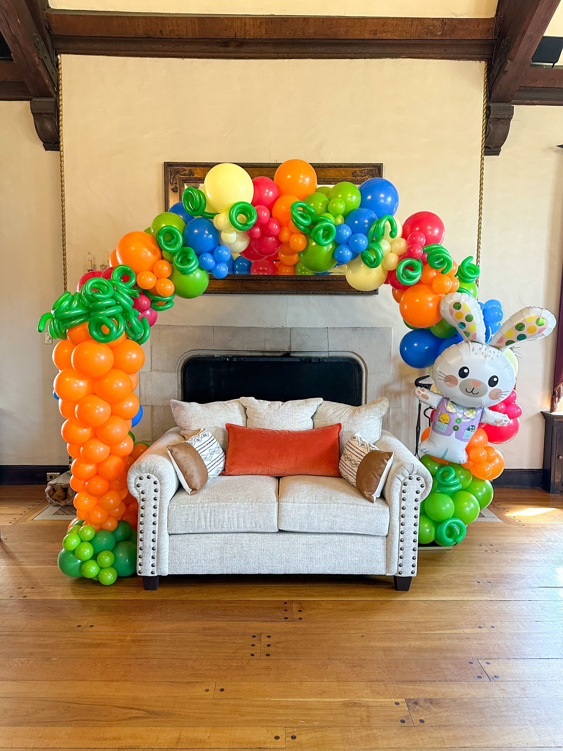 Easter-themed balloon arch with carrot, bunny, and colorful balloons over a couch in a room.
