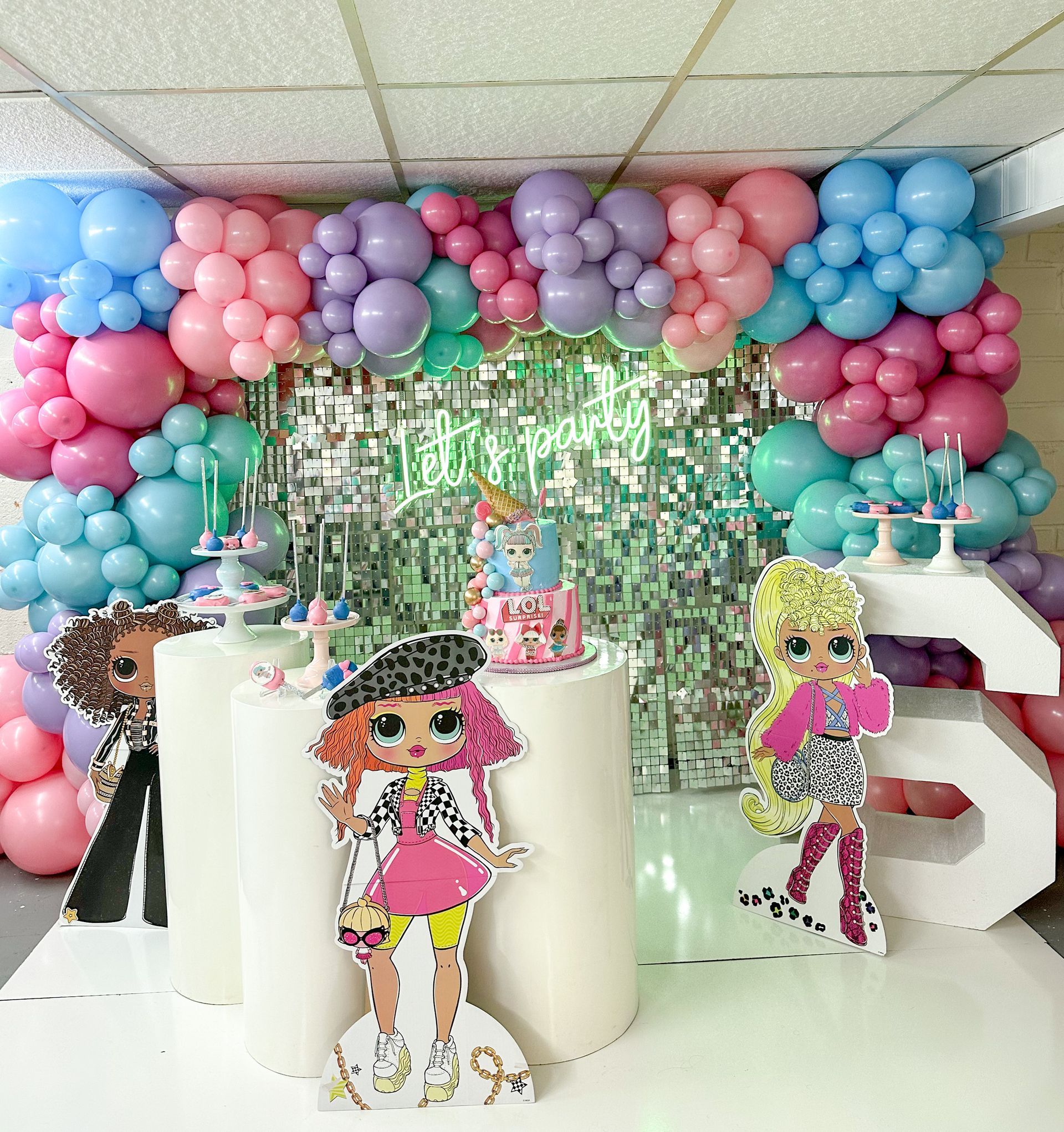 Party backdrop with balloons, a cake, and LOL Surprise dolls. Pastel colors.