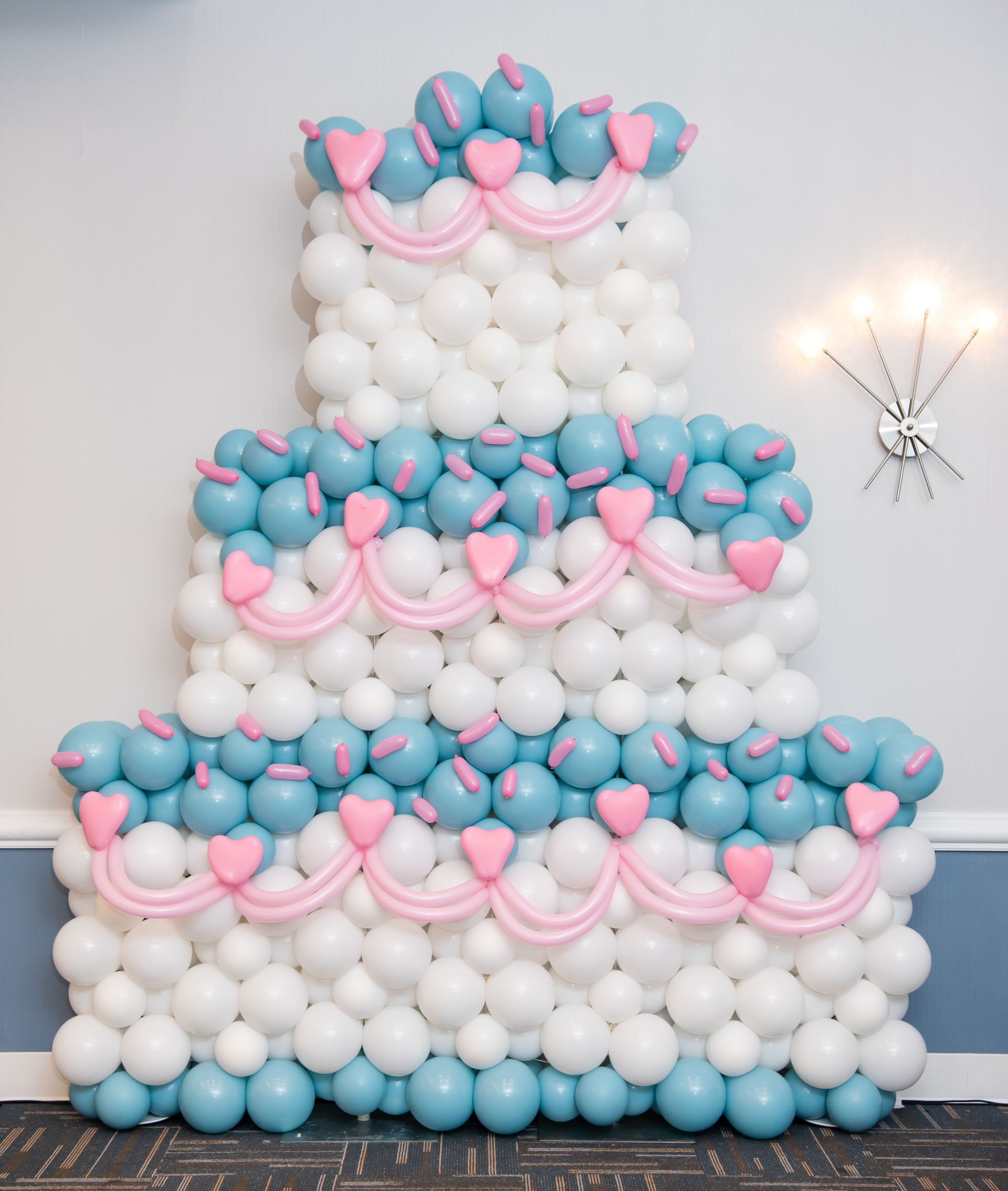 Three-tiered balloon cake with white, blue, and pink decorations against a wall.