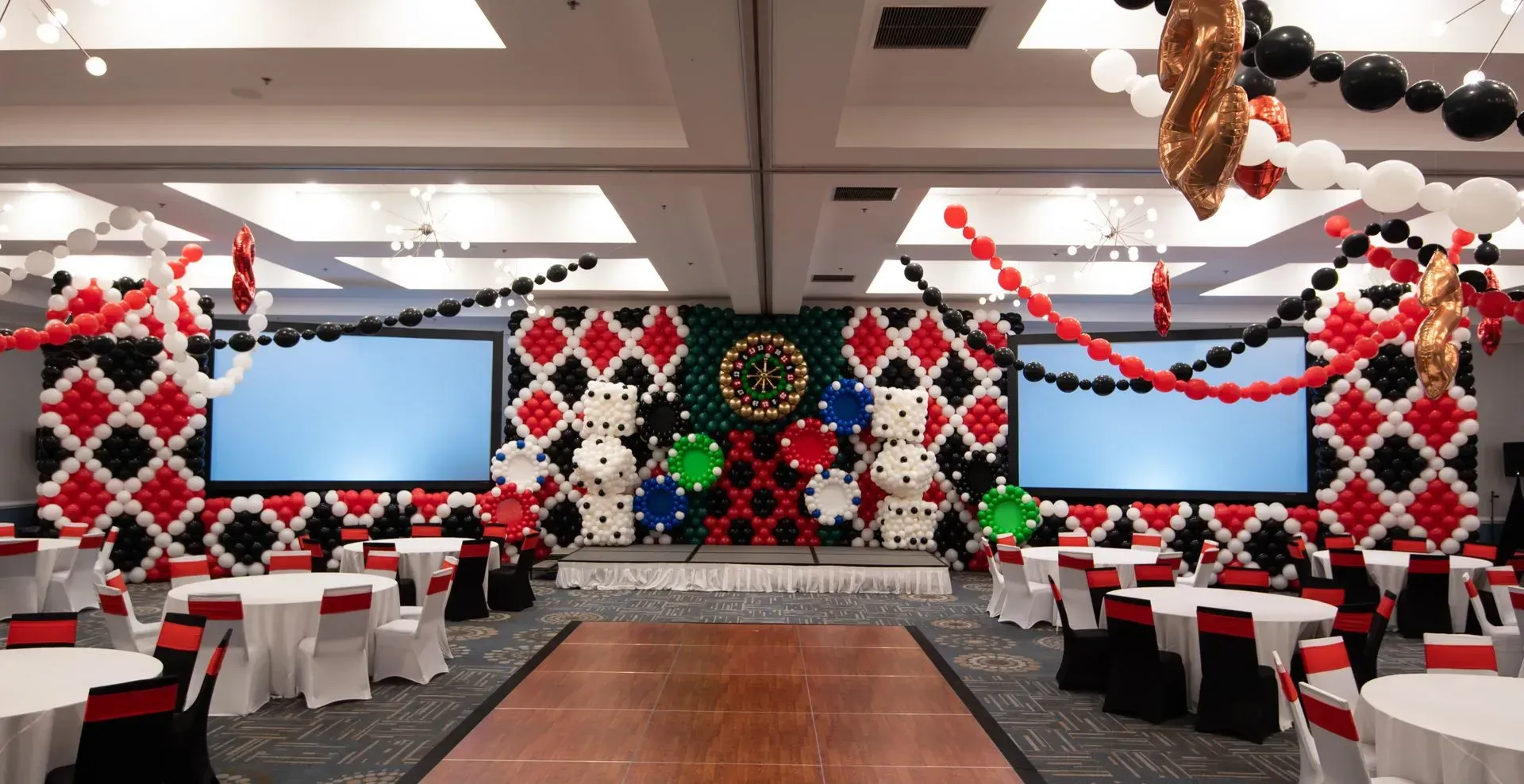 Casino-themed event hall with tables, stage featuring large dice, playing card patterns, and balloons.