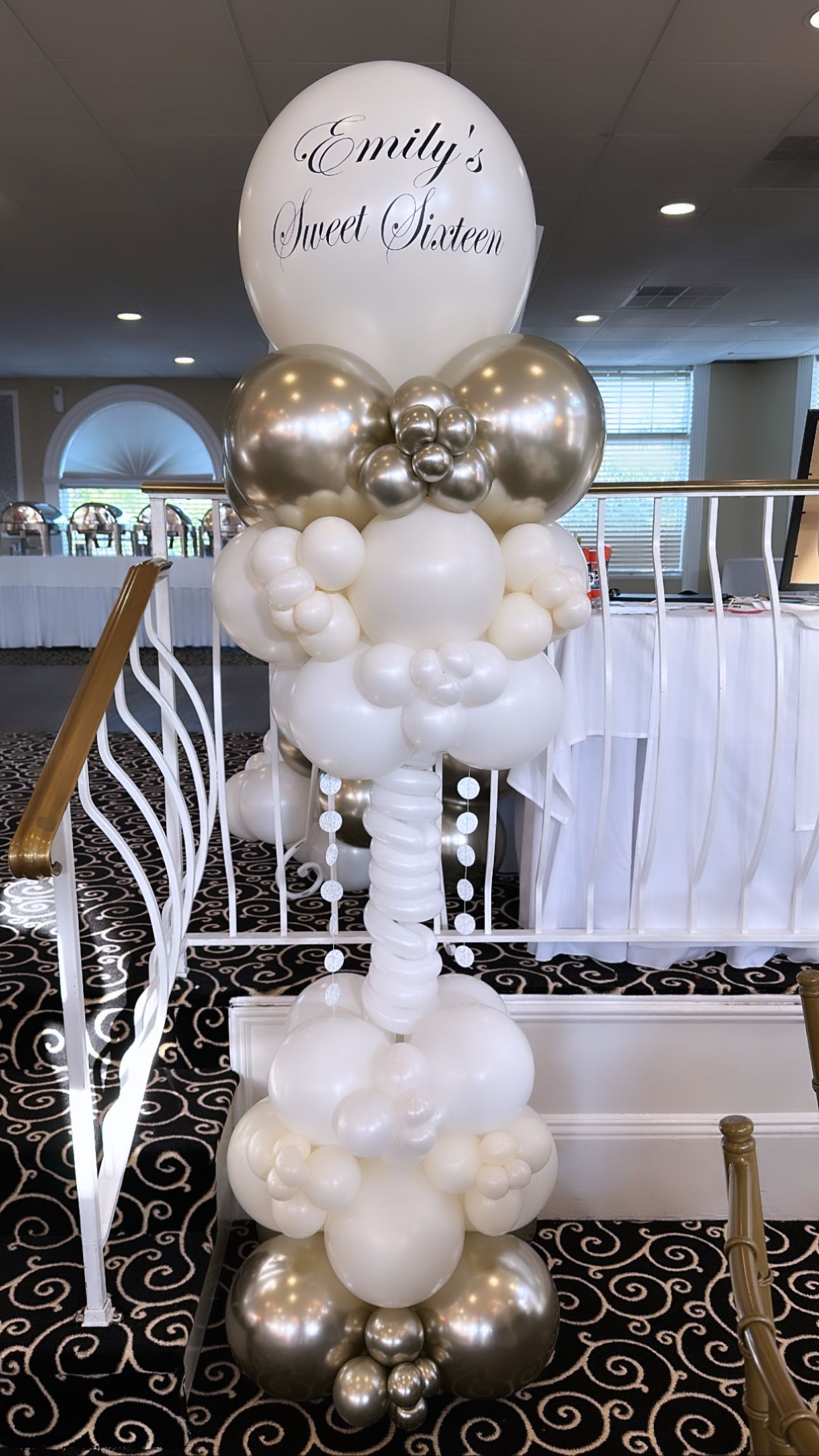White and gold balloon column with a round sign in a decorated indoor event venue