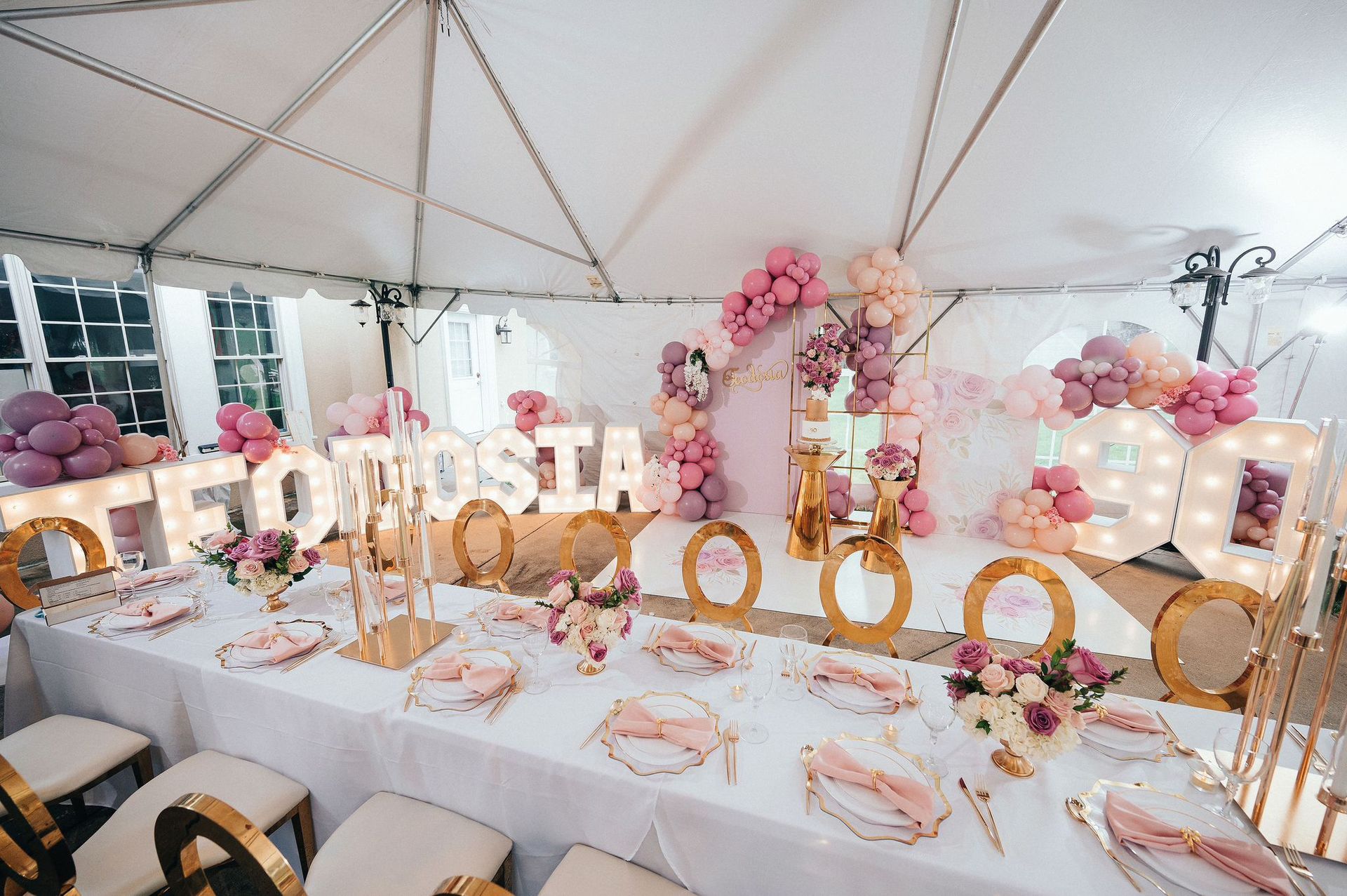 Pastel balloon-decorated party table under a white tent with gold letters and floral accents
