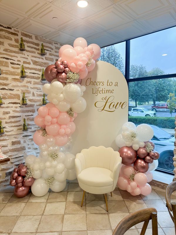 Pastel balloon arch beside cream chair and “There’s a Lifetime of Love” backdrop in a bright venue