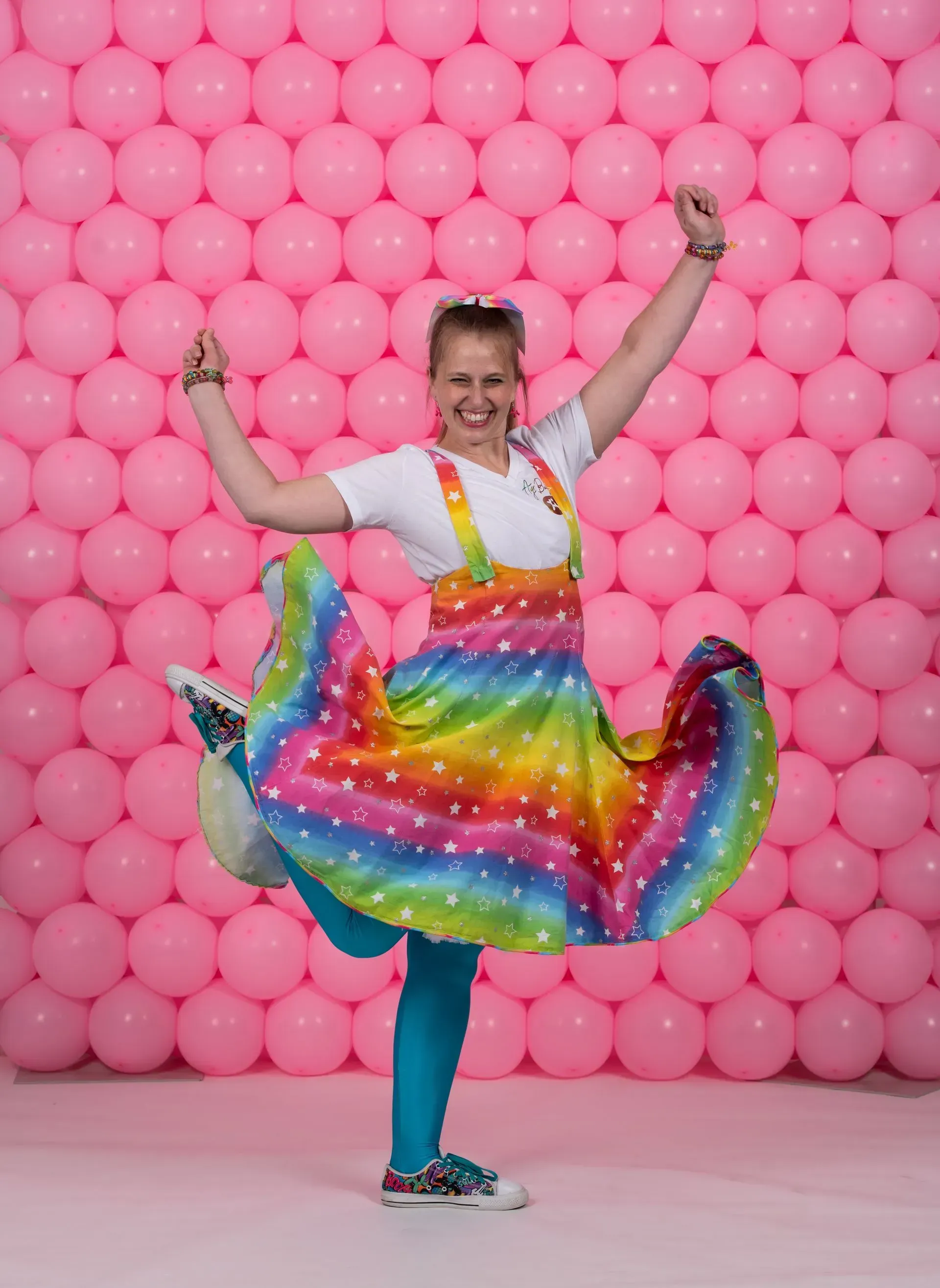 Person in rainbow skirt and suspenders dances in front of pink balloon wall, arms raised, smiling.