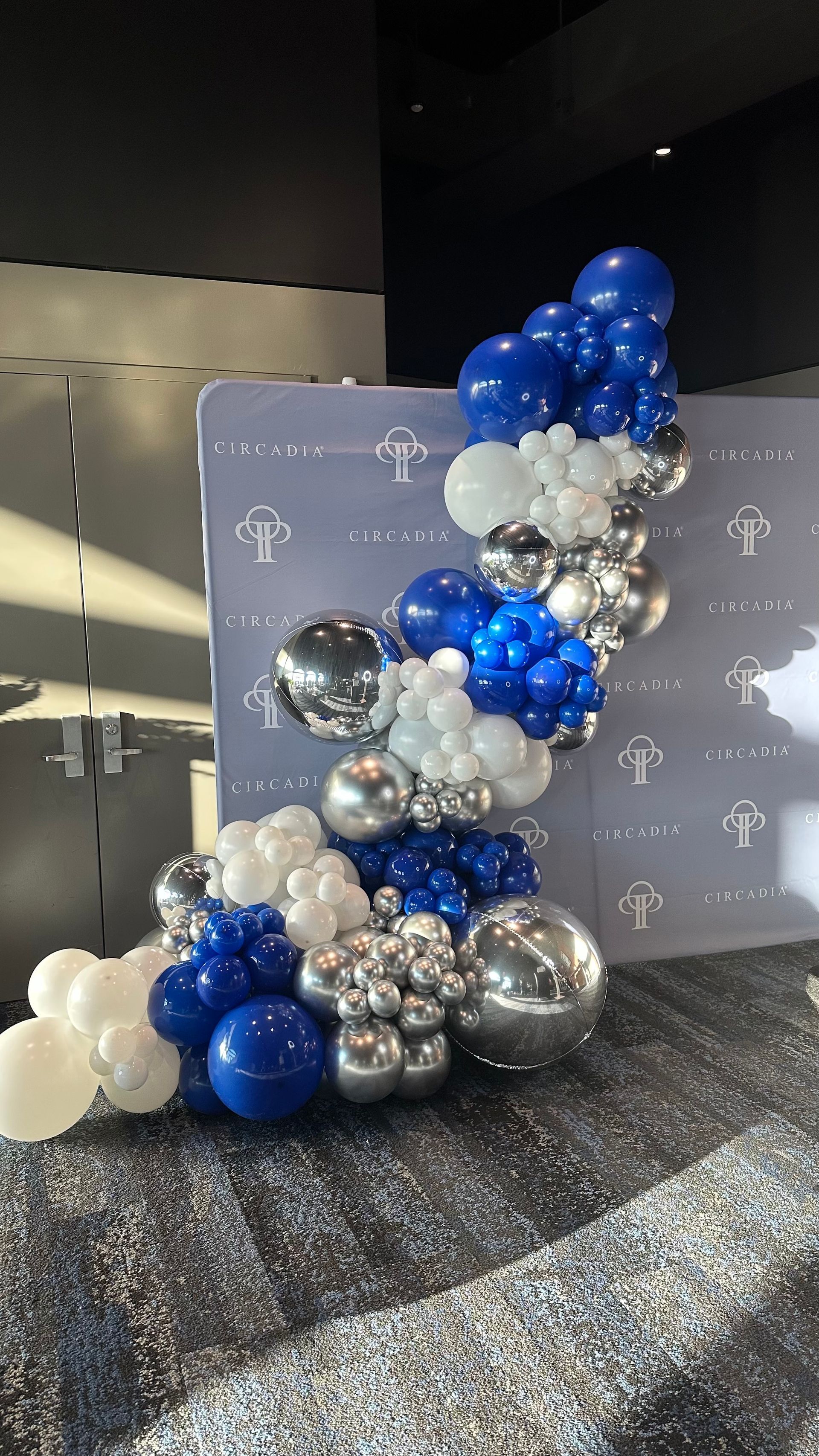 Blue, white, and silver balloon arch beside a gray event backdrop on a glittery floor