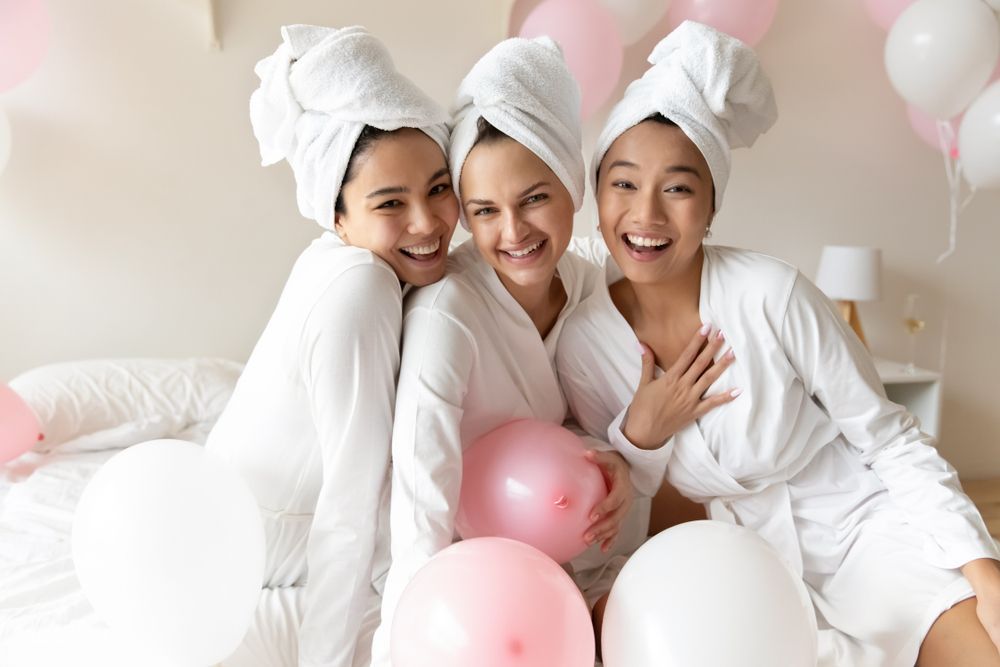 Three smiling friends in white robes with towels on their heads, seated among pink and white balloons.