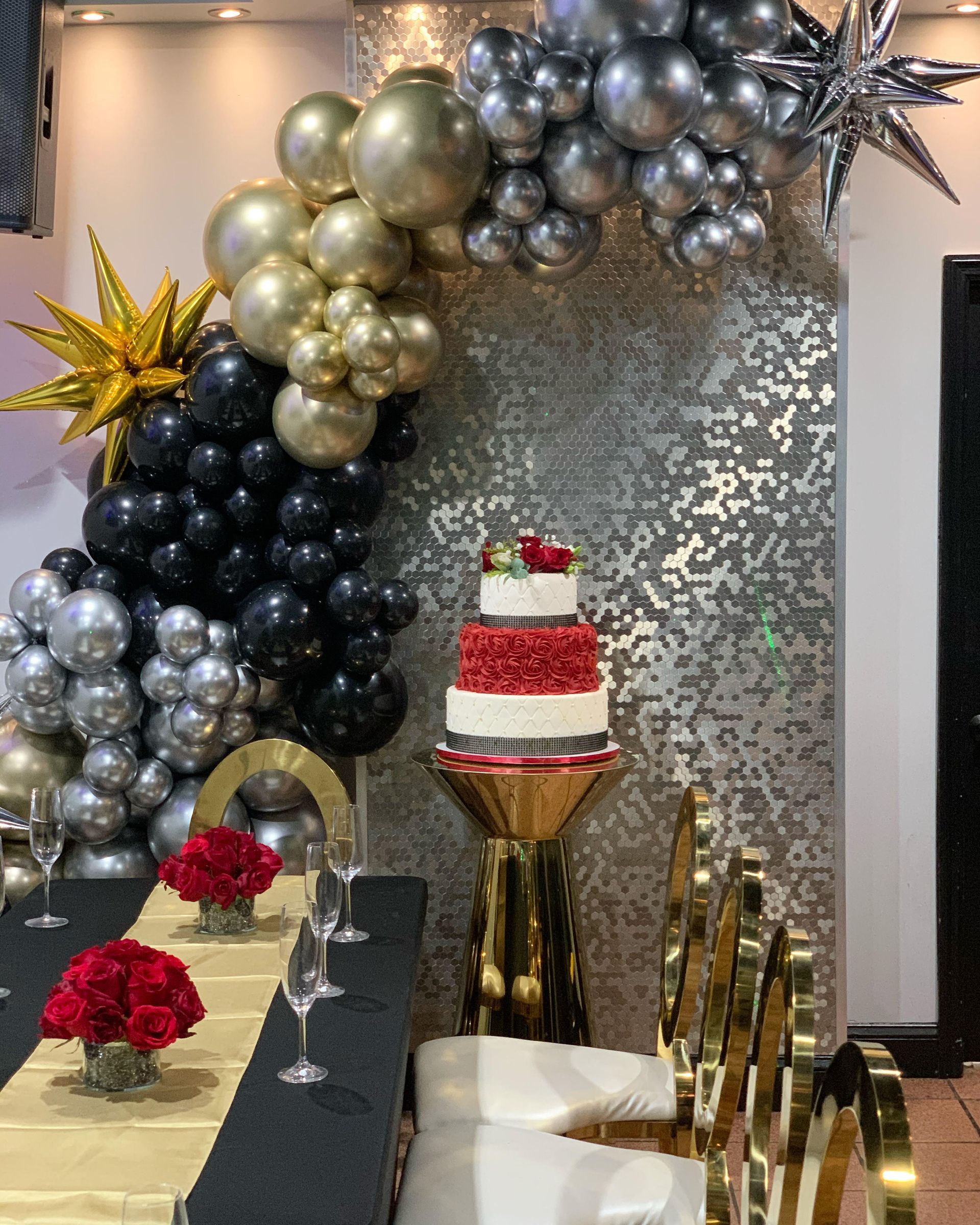 Decorated dessert table with black, silver, and gold balloons and a tiered red-and-white cake