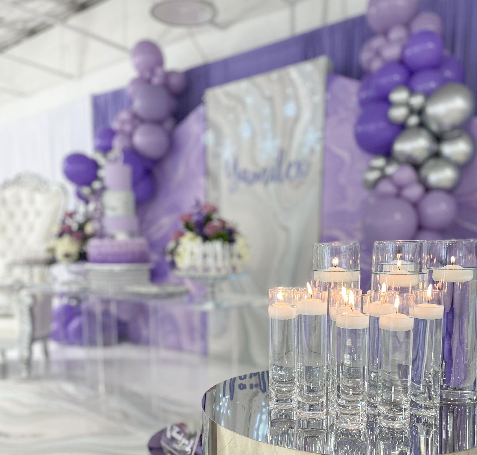 Lavender and silver event decor with balloons, floral arrangements, and candlelit glass cylinders on a reflective table