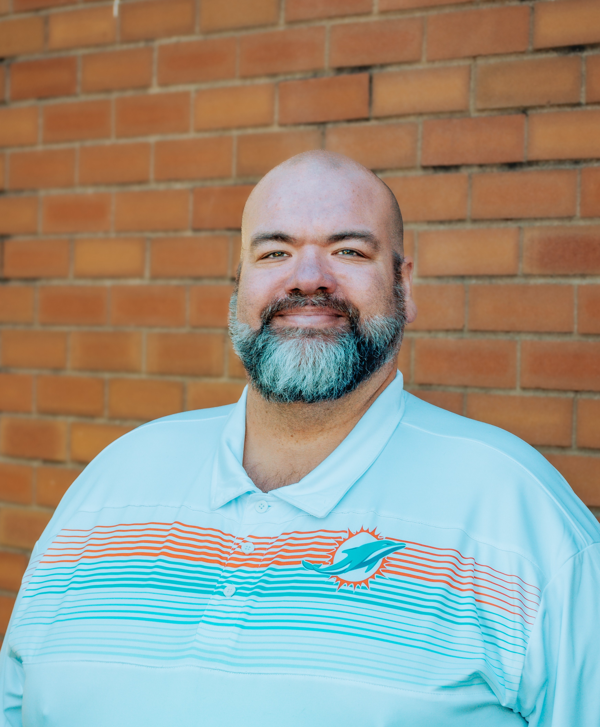 A man smiling in front of a brick wall wearing a dolphin logo polo