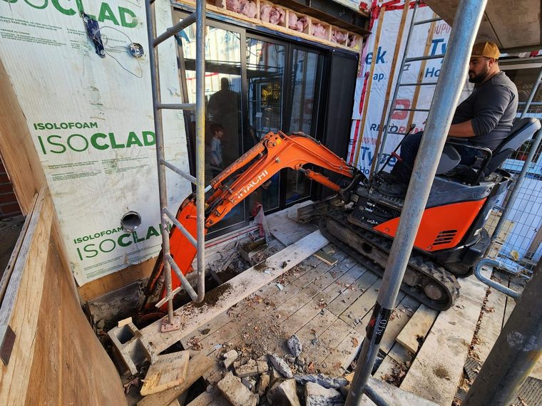 Une mini-pelle orange travaille sur un chantier de construction, à côté de murs à ossature bois et d'un opérateur assis.