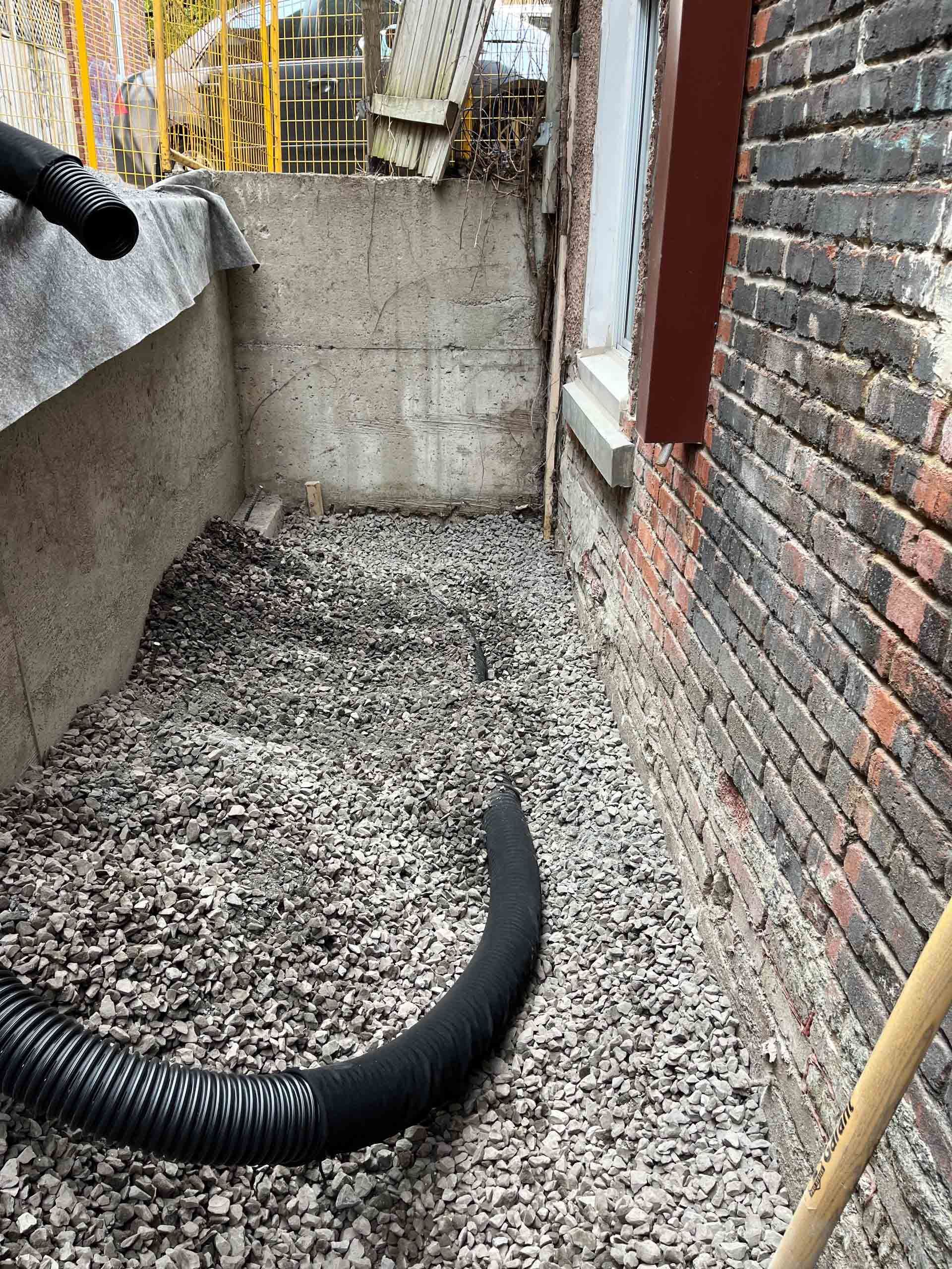 Allée étroite en gravier dans une cour arrière, avec des murs en briques et un tuyau d'arrosage noir ondulé au sol.