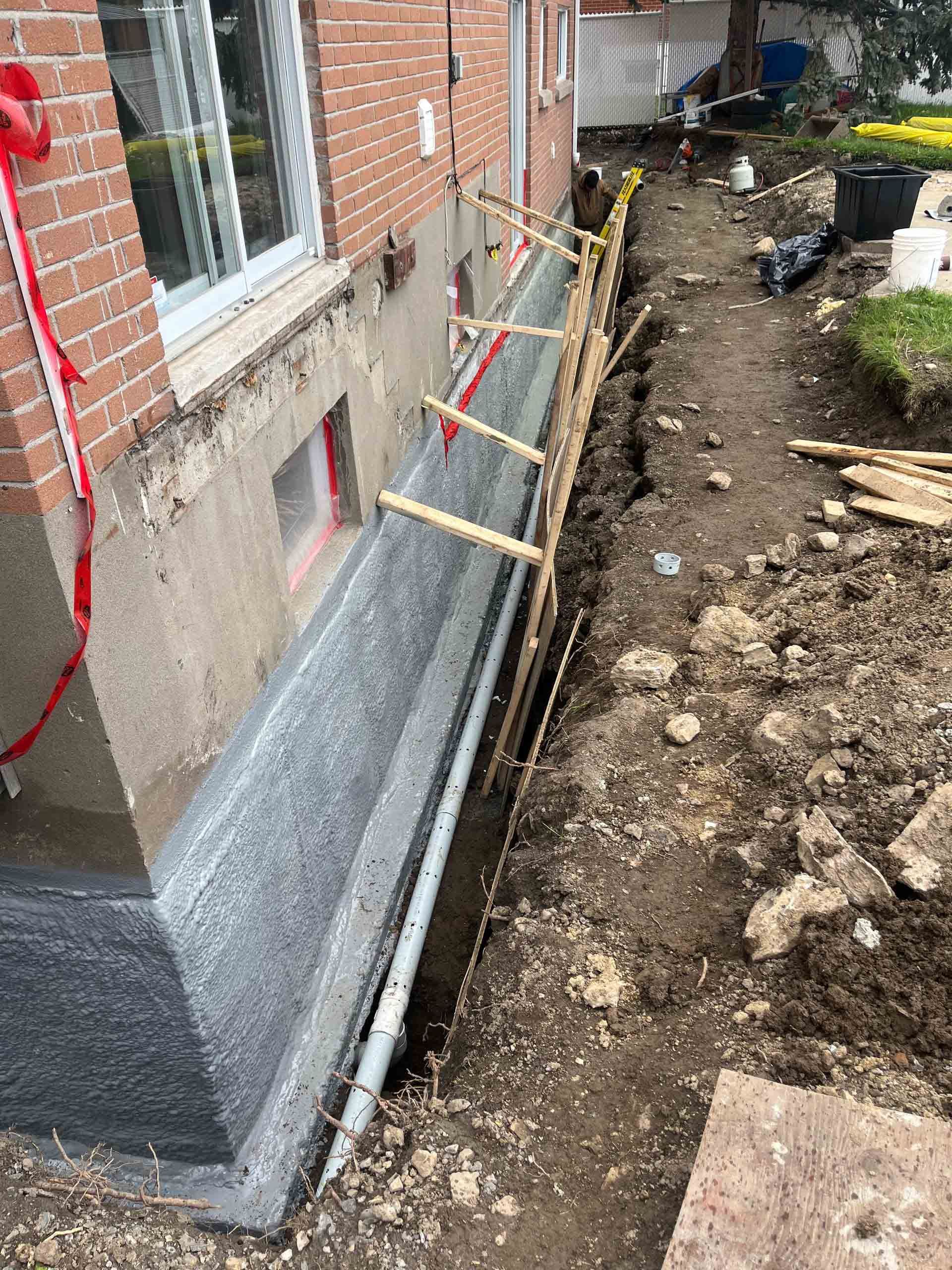 Tranchée de construction le long des fondations d'une maison en briques, avec poutres de soutien et isolation murale apparente.