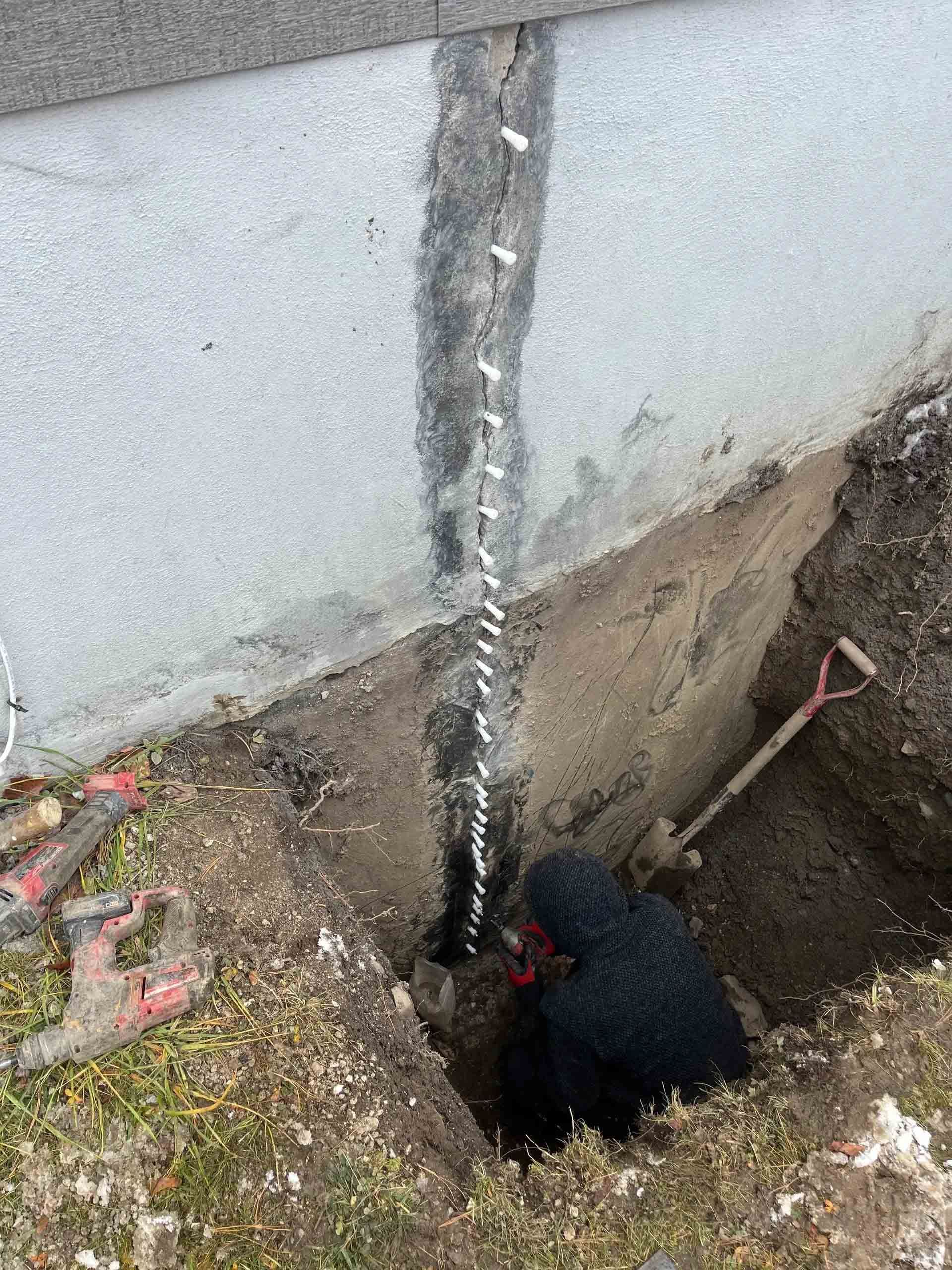 Main gantée de noir travaillant dans une tranchée de terre à côté d'un mur de bâtiment présentant une fissure verticale ou un tuyau.