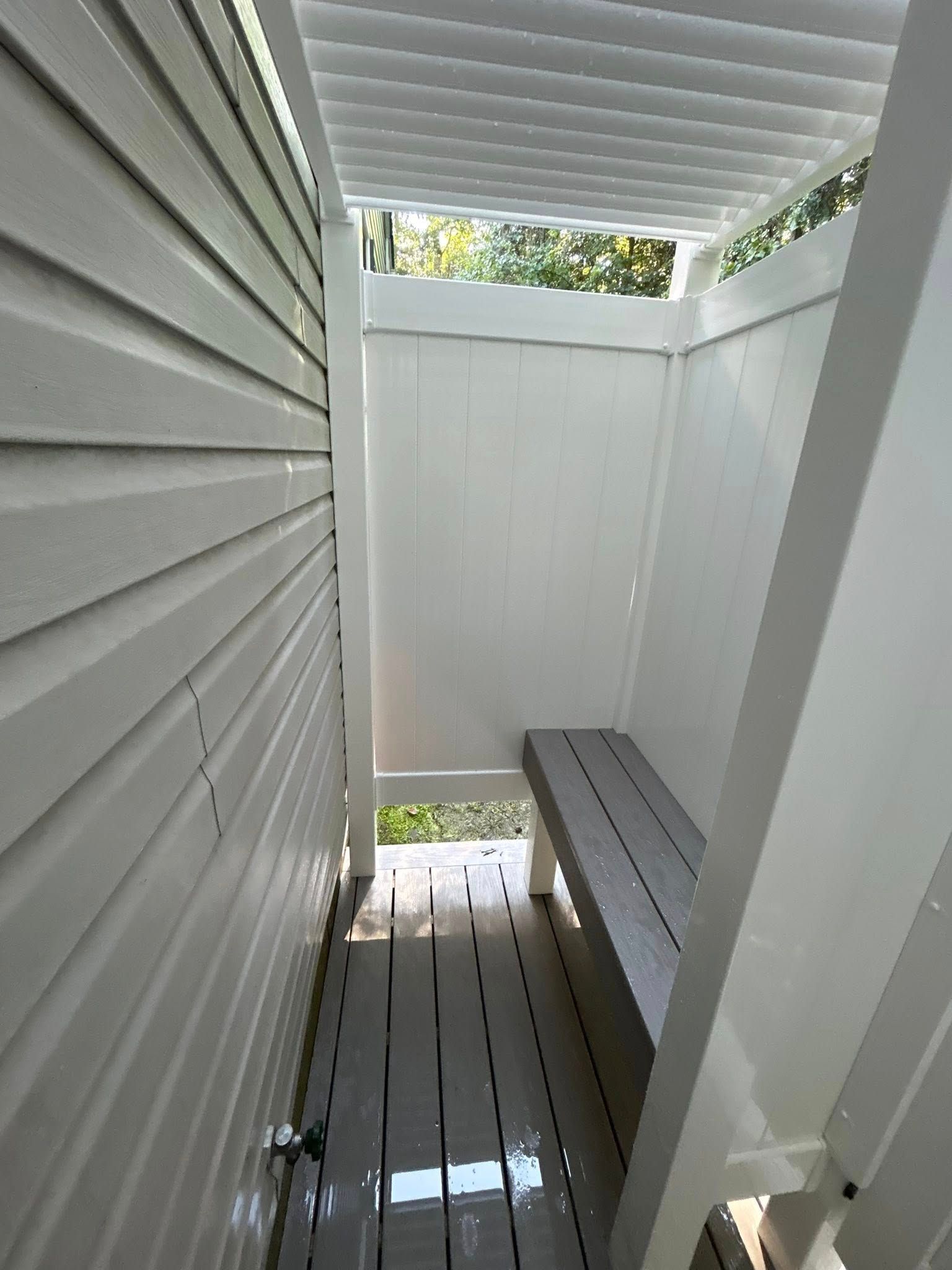 Narrow outdoor shower with white siding, bench, and wet floor beneath a slatted roof.