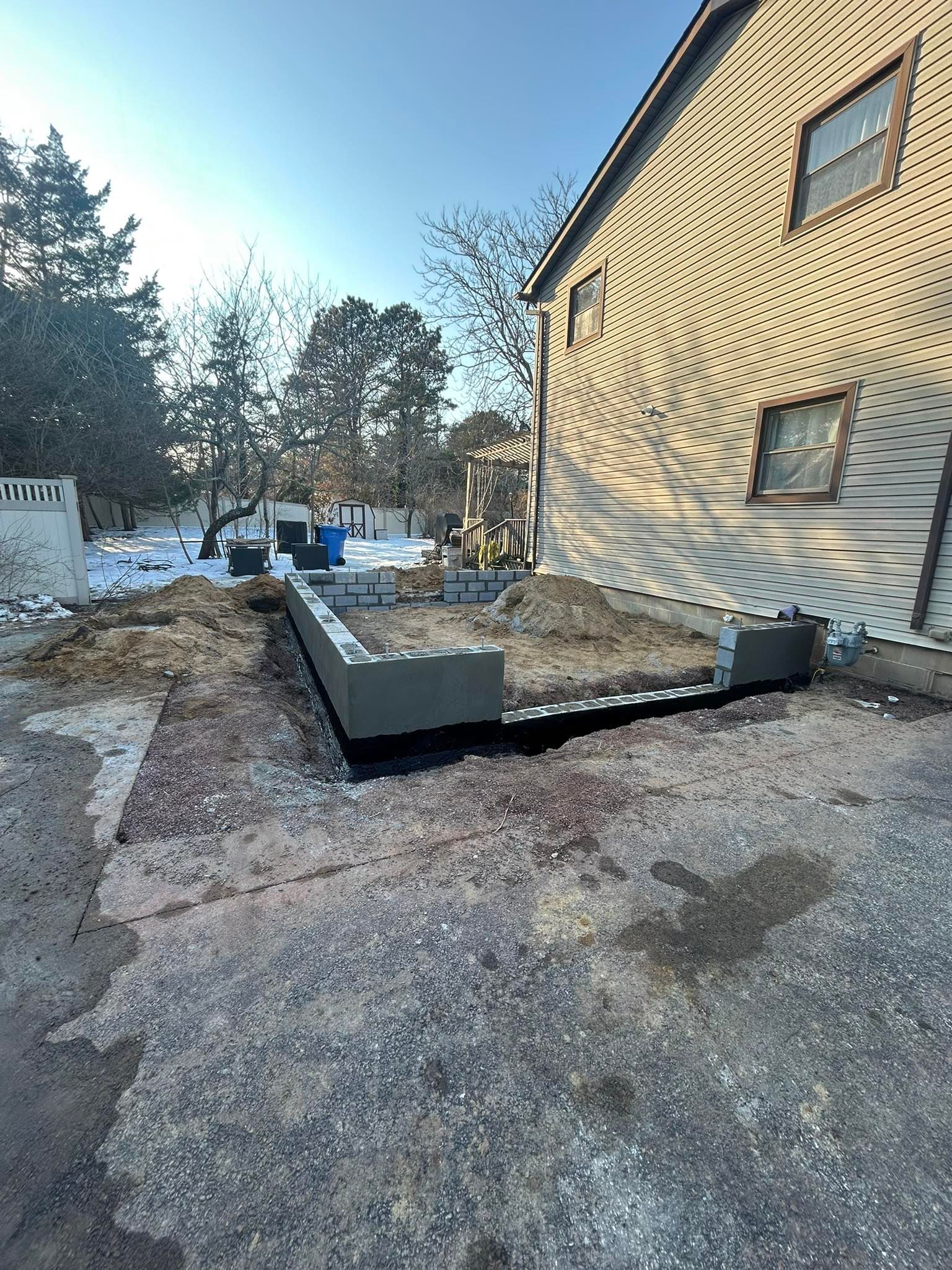 Concrete foundation footing beside a house in a snowy yard, with trees in the background.
