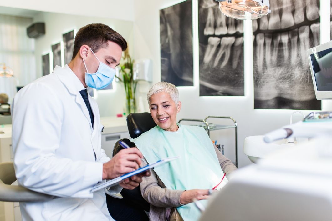 Dentist with mask consults a smiling patient in a dental chair, taking notes. X-rays on wall.