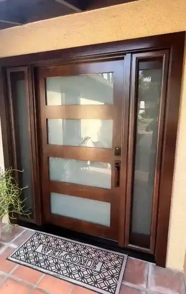 Modern wooden front door with frosted glass panels and a narrow side window, viewed from outside with a doormat.