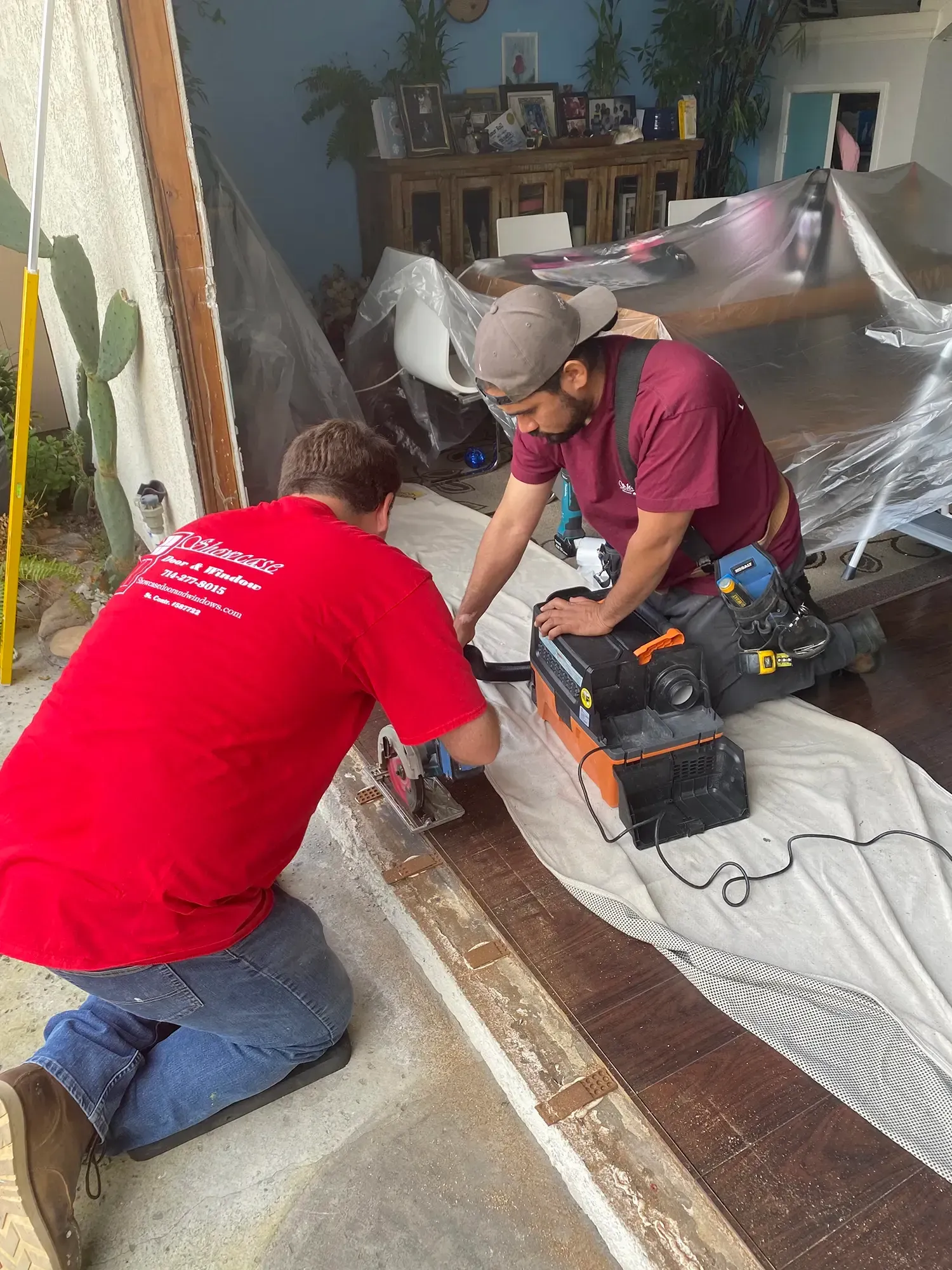 Two workers kneel on a floor, using power tools to repair a wooden threshold under plastic sheeting.