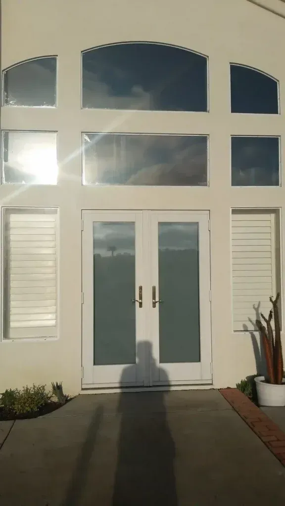 Sunlit white house front entrance with double glass doors, windows, and potted plants on either side