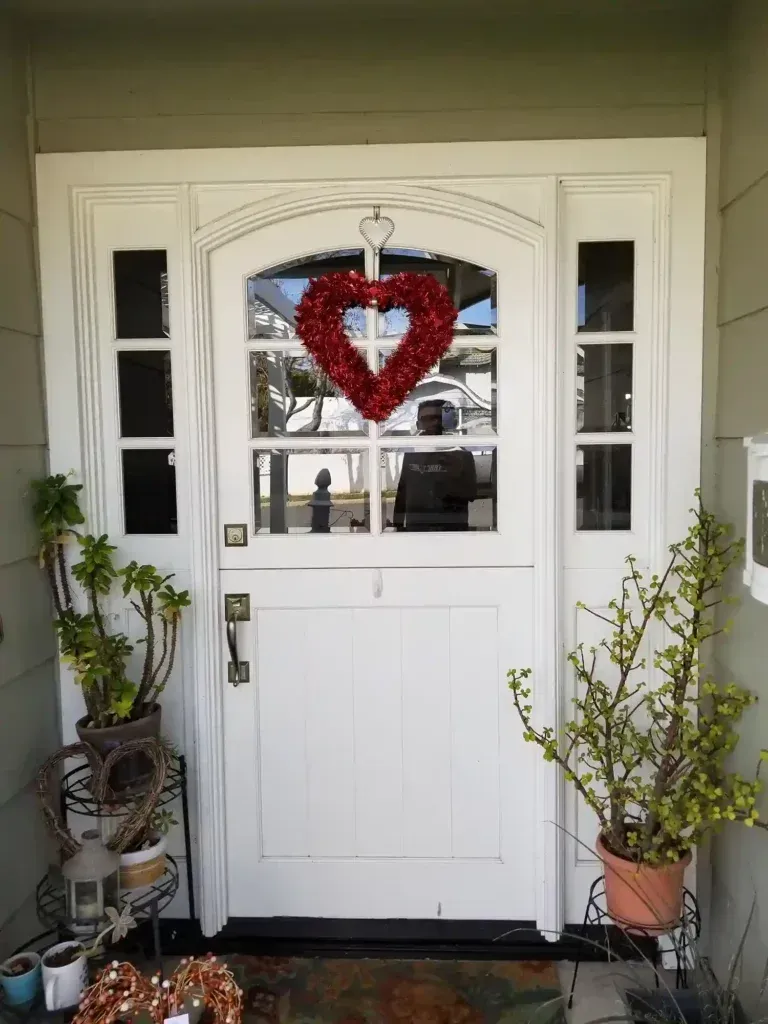 Front porch with white door, potted plants, and a red heart wreath hanging in the window.