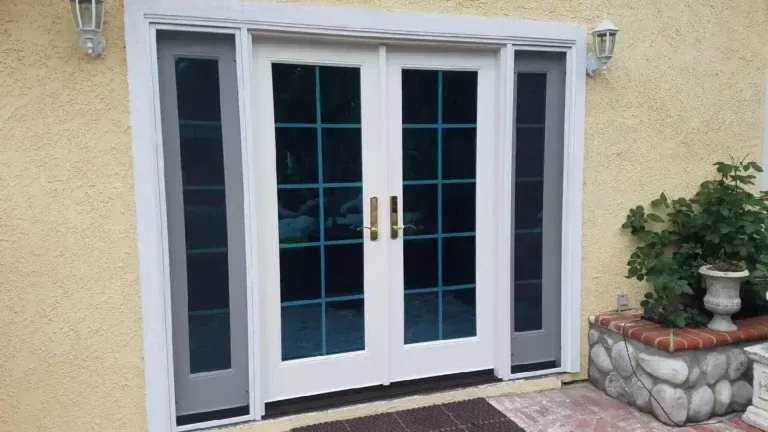 Front entrance with white double doors and sidelights on a yellow stucco house, next to a planter and small patio.