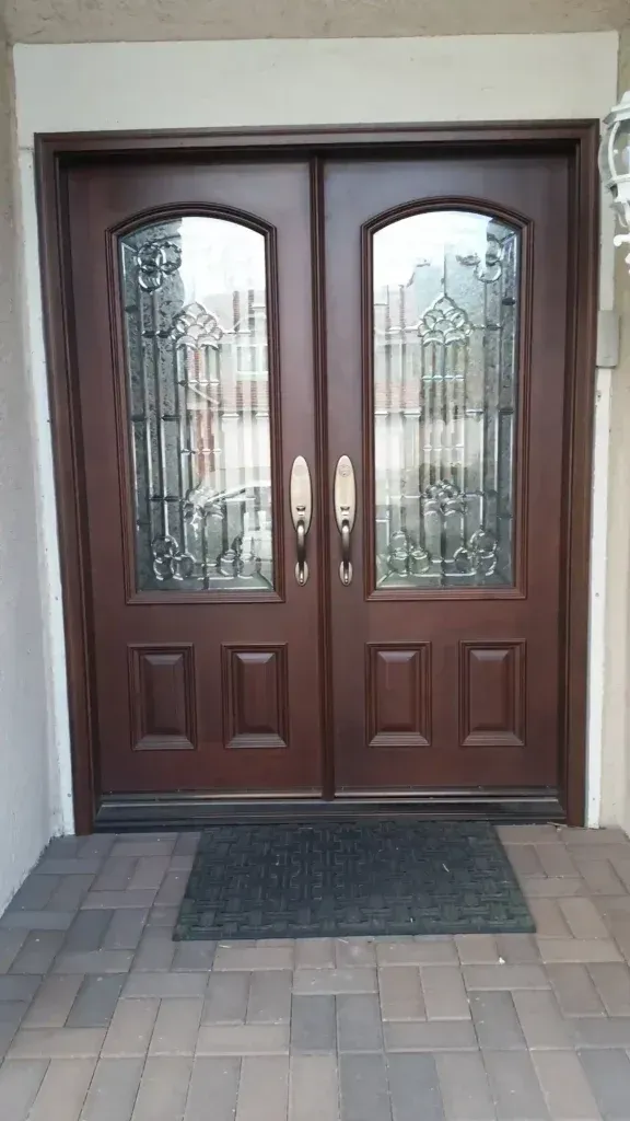 Brown double front doors with glass panels and brass handles, set in a stone entryway with a doormat.