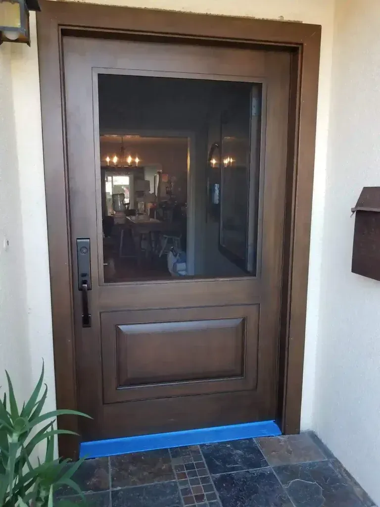 Wooden front door with glass panel and black handle, framed by white stucco wall and blue threshold tape