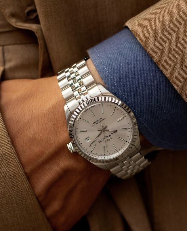 Orologio d'argento al polso; al braccio una camicia blu con un blazer marrone chiaro.