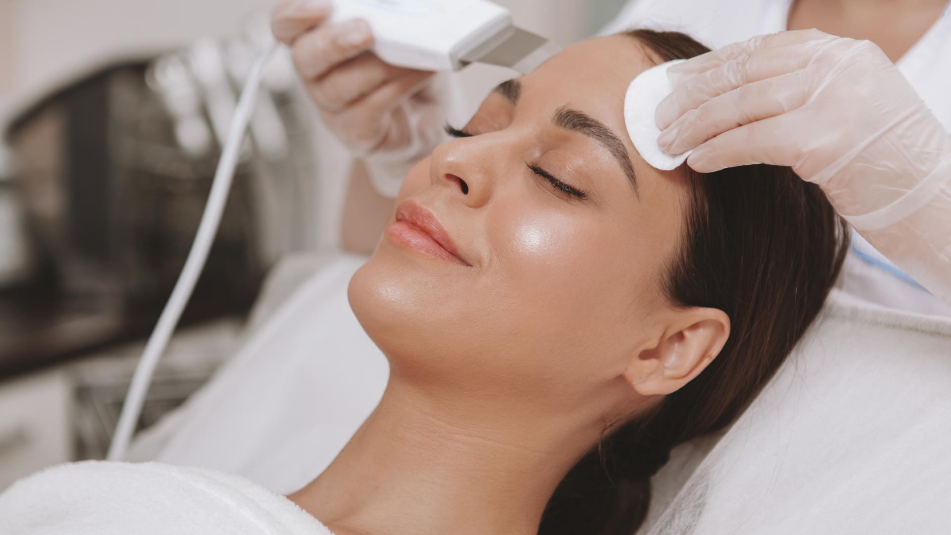 Woman receiving a facial treatment while lying on a spa bed, with gloved hands applying skincare to her face