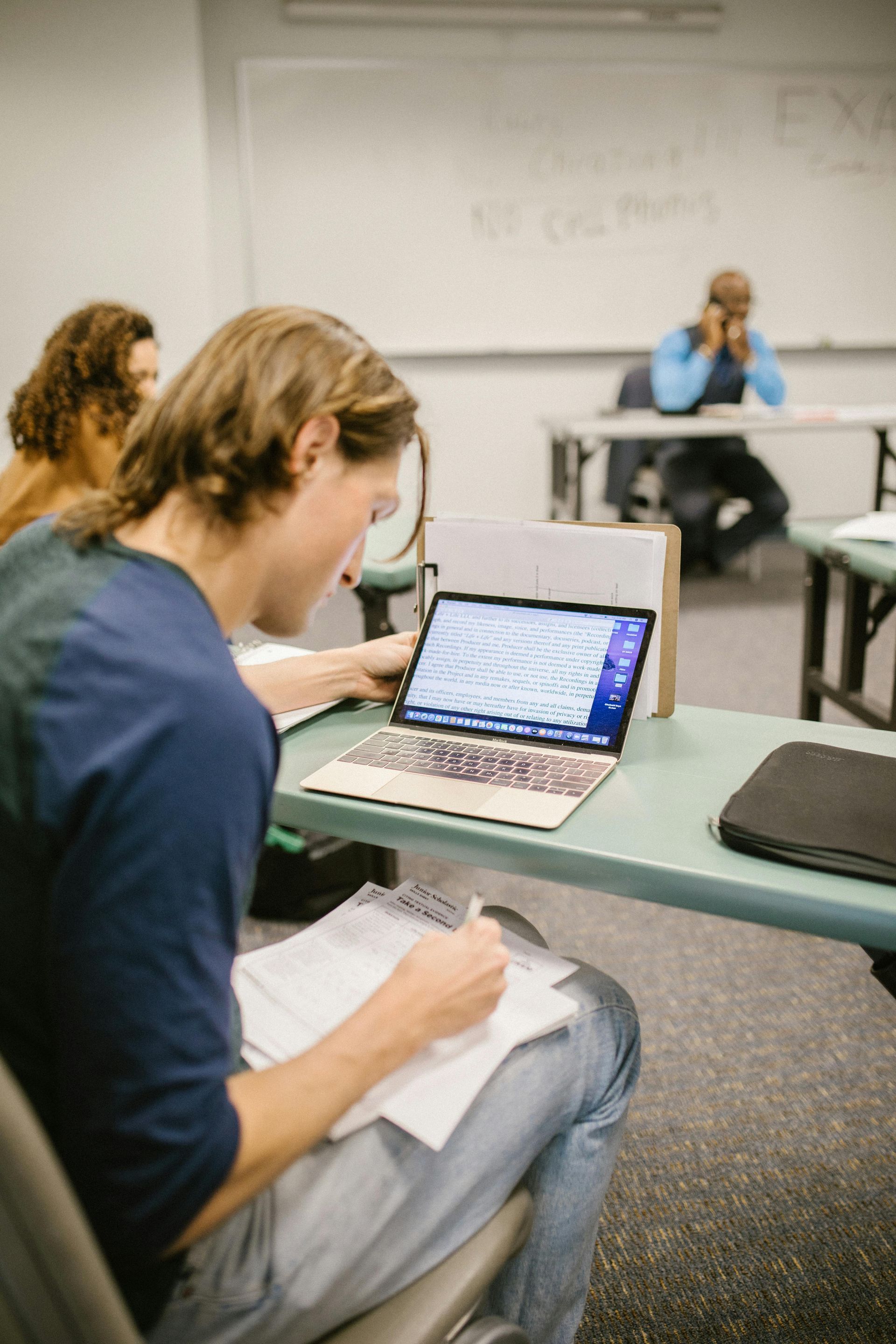 Student in a classroom using a laptop and taking notes. Another student appears in the background.