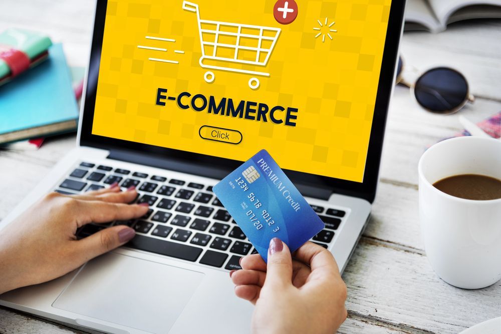 Hands holding a credit card over a laptop displaying “E-COMMERCE” on a yellow screen.