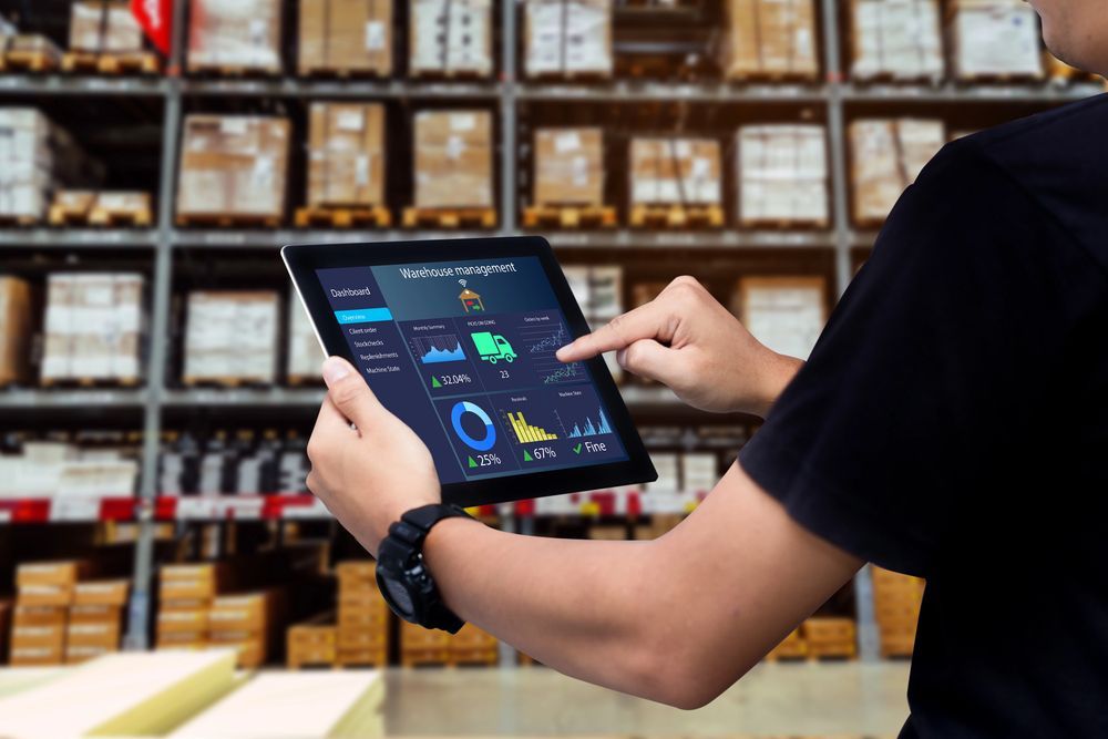 Person using a tablet in a warehouse aisle with stacked boxes in the background