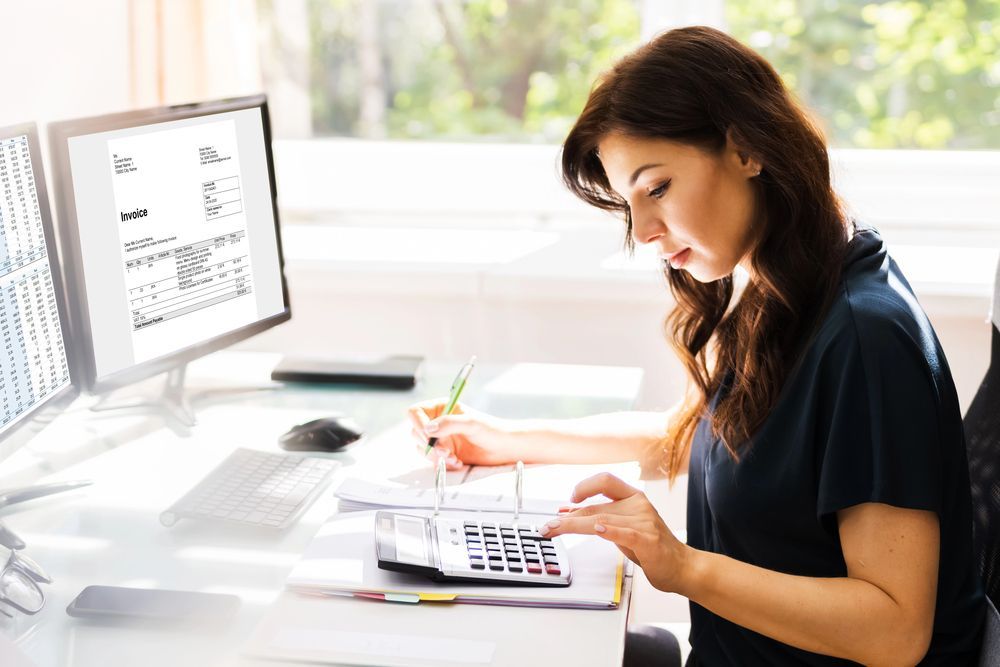 Woman working at a desk with a computer and calculator, reviewing paperwork in a bright office