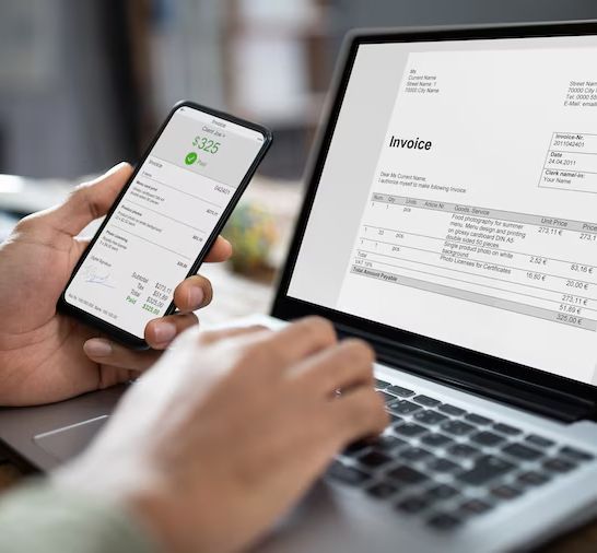 Person using a smartphone and laptop to review an invoice on a desk