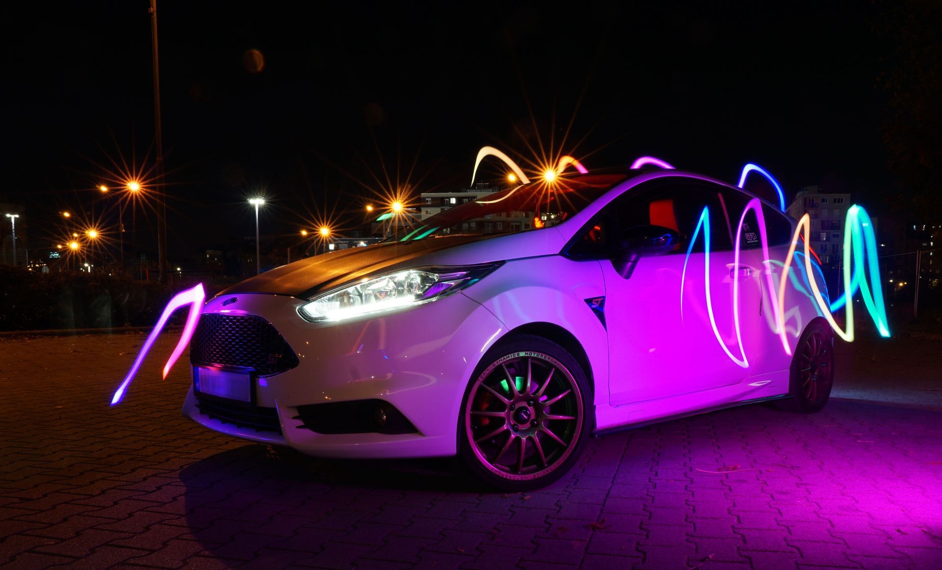 A white Ford Fiesta ST hatchback parked at night on a paved surface, illuminated by vibrant, multi-colored light painting effects. Neon-like waves of pink, blue, orange, and green light wrap around the body of the car, with the driver's side glowing in a distinct bright purple hue. The vehicle's headlights are turned on, and multiple warm streetlights glow in the dark background.