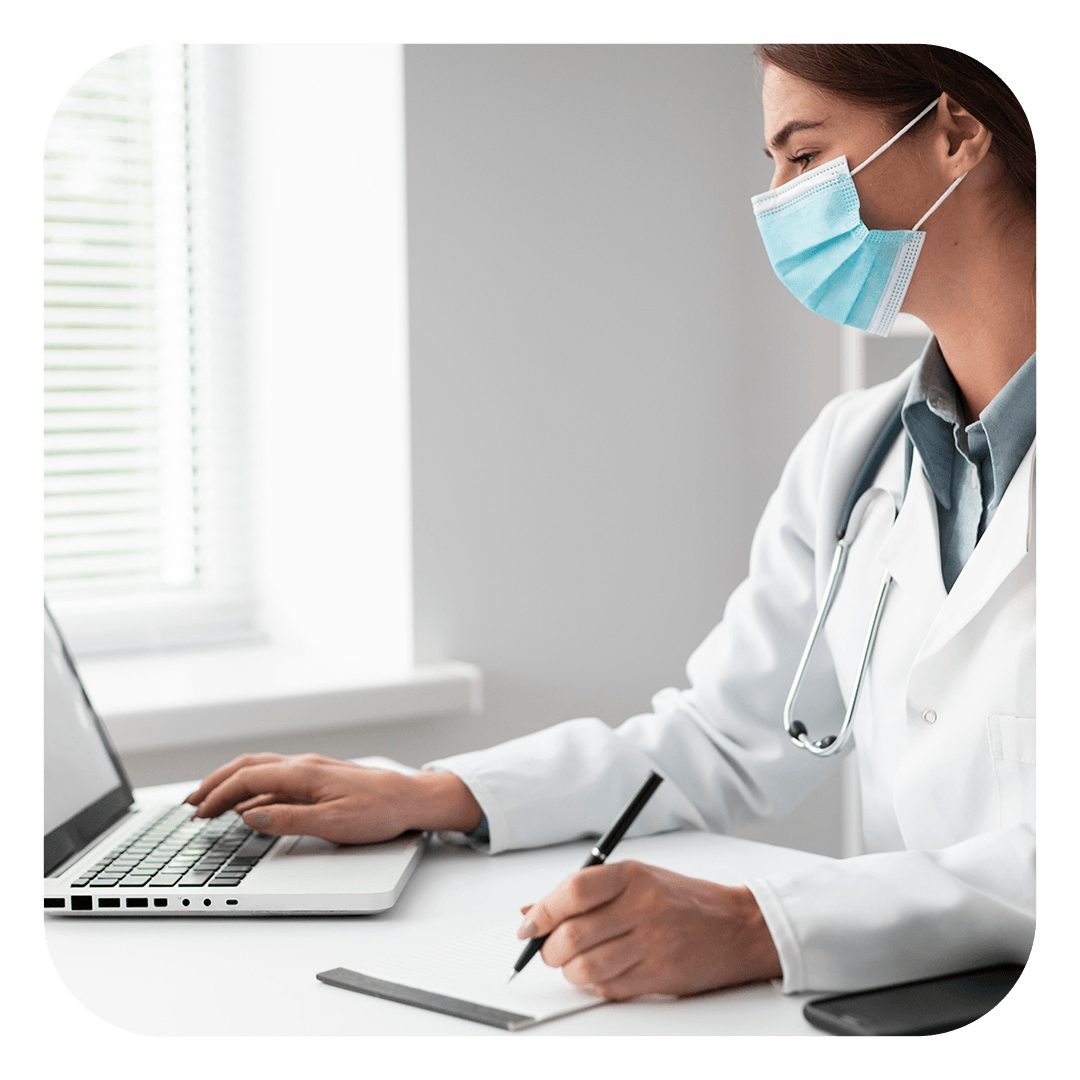 Um profissional da área médica, vestindo jaleco branco e máscara facial azul, digita em um laptop enquanto faz anotações em uma mesa.